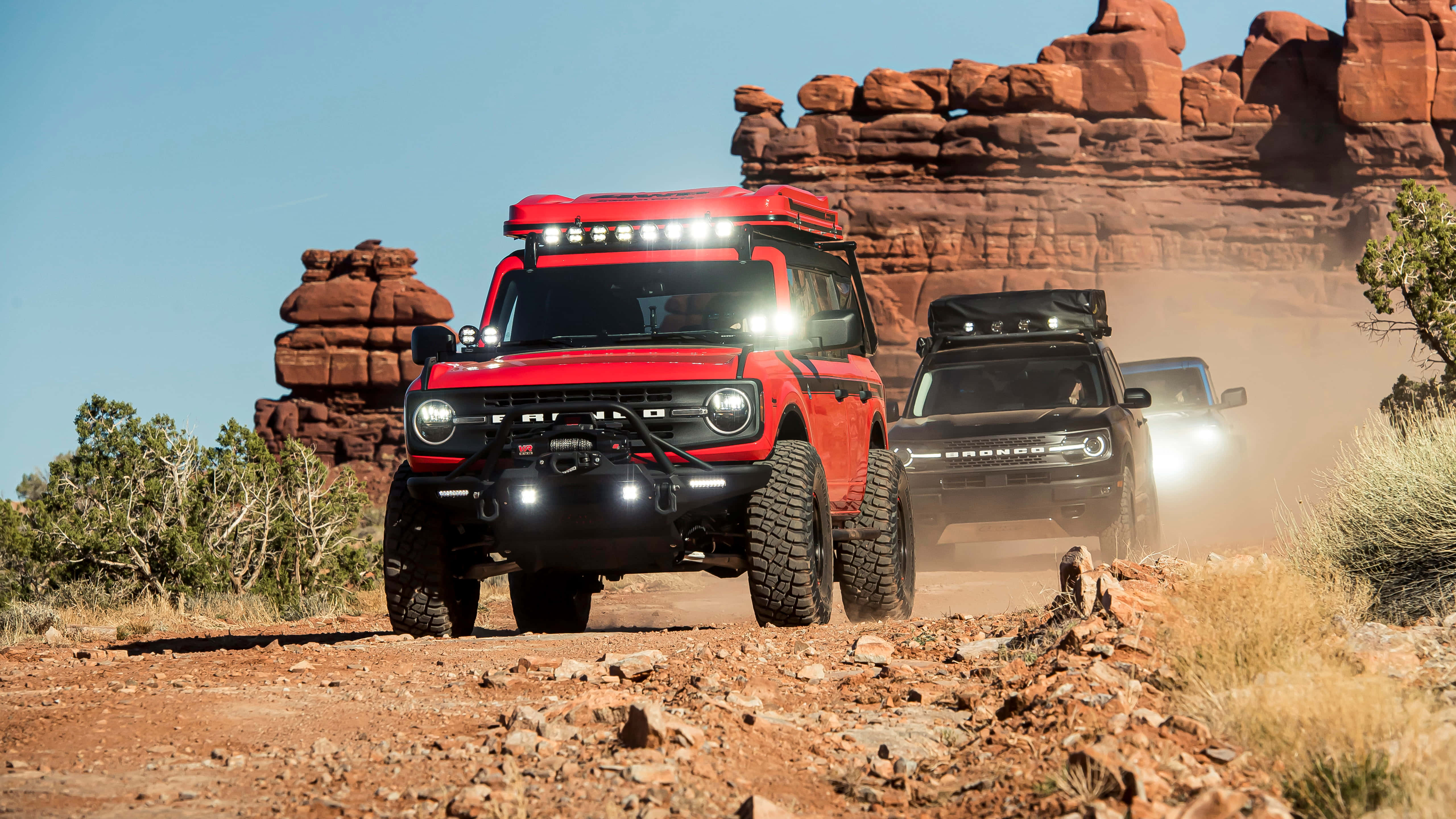 Ford Bronco 桌布 壁紙 — 兩輛堅固的越野車在晴朗的藍天下，在雄偉的紅色岩層下的塵土飛揚的沙漠小徑上疾馳。