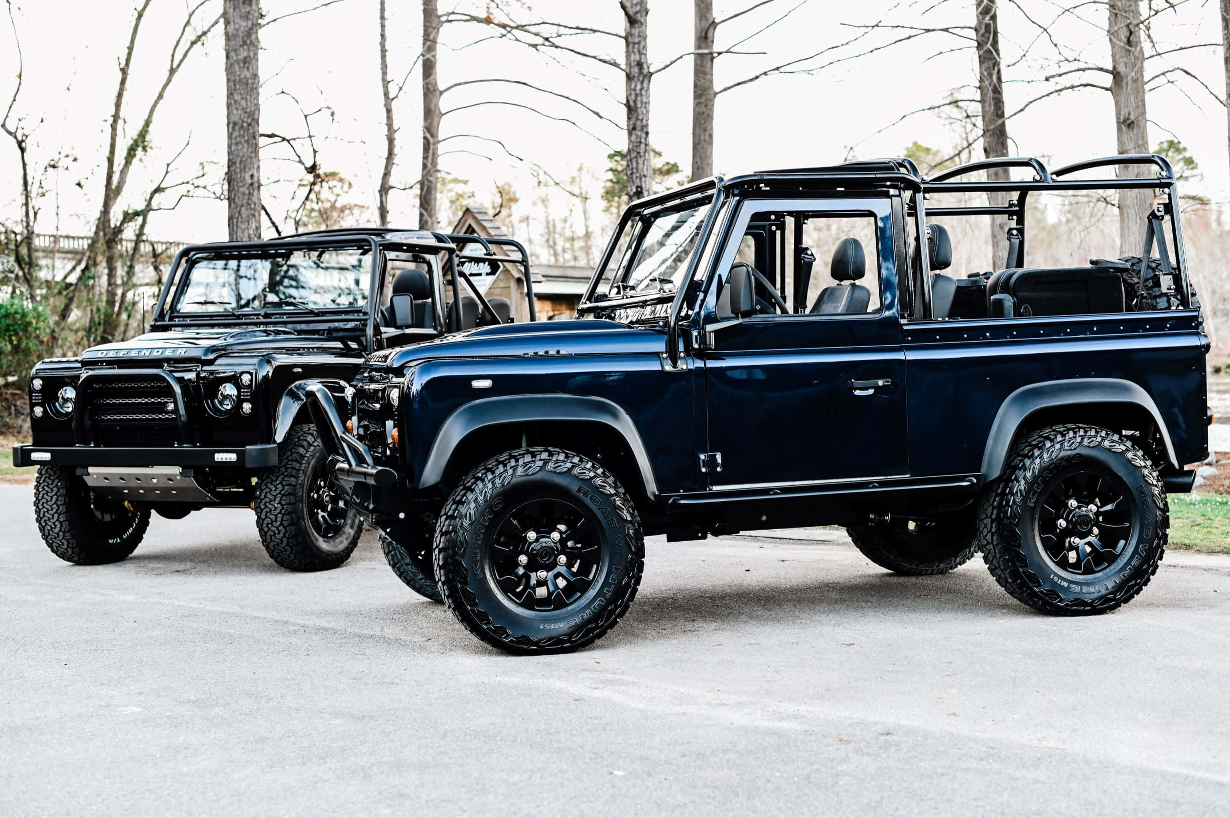 Land Rover Defender 桌布 壁紙 — 兩輛堅固的黑色 Land Rover Defender 並排停放在鋪砌路面上，背景有光禿禿的樹木。