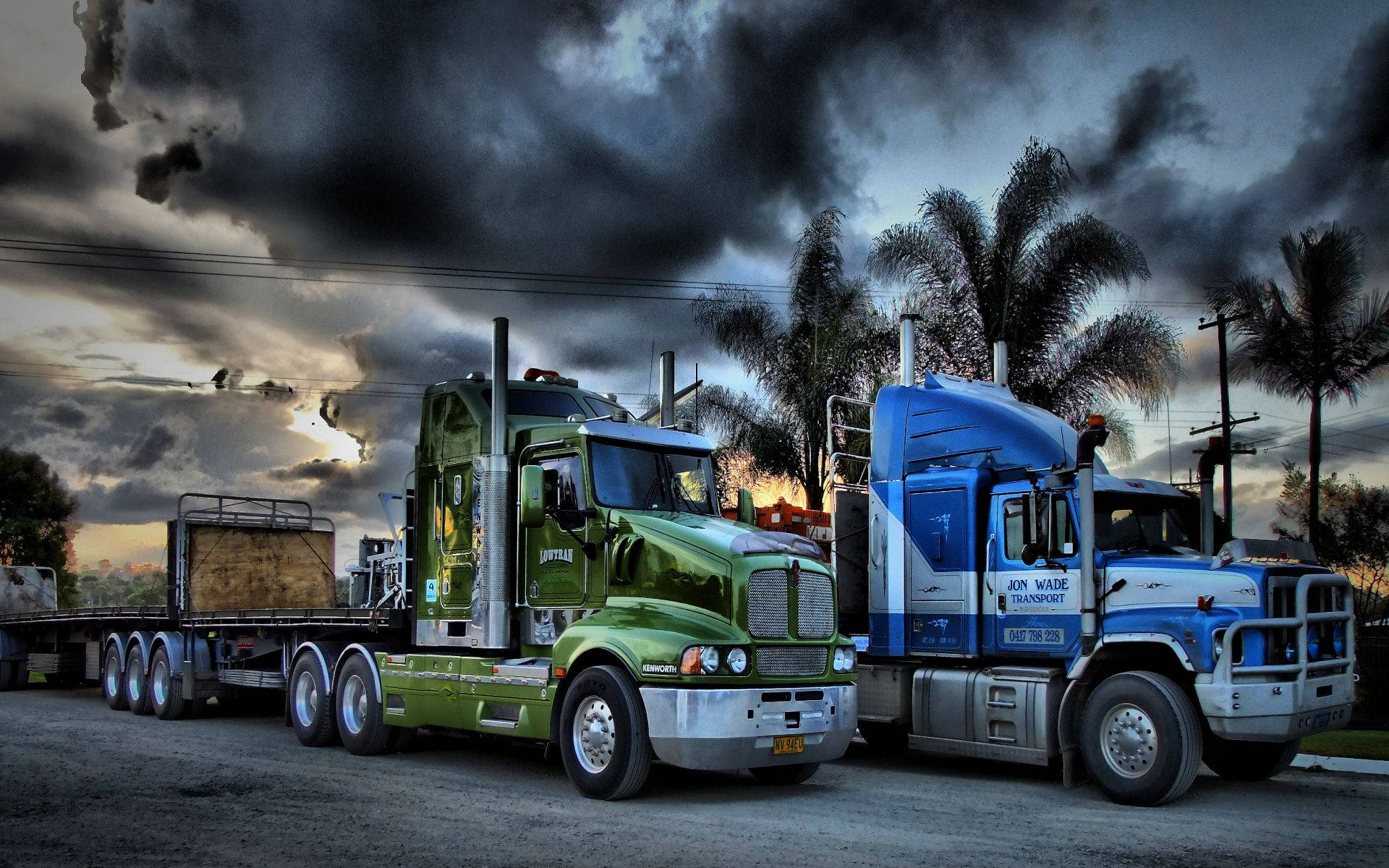 Trucks 桌布 壁紙 — 兩輛大型半掛卡車，一綠一藍，並排停放在戲劇性的暴風雲下，背景有棕櫚樹。