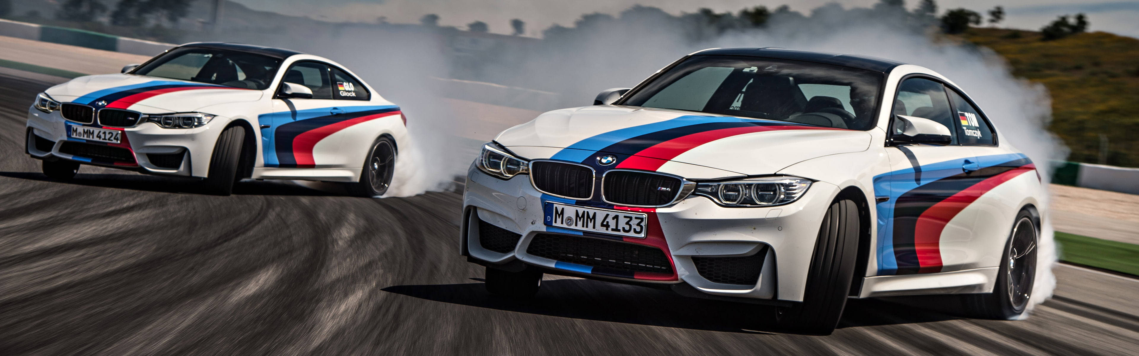 兩輛BMW M4賽車在賽道上甩尾，身後留下煙霧。