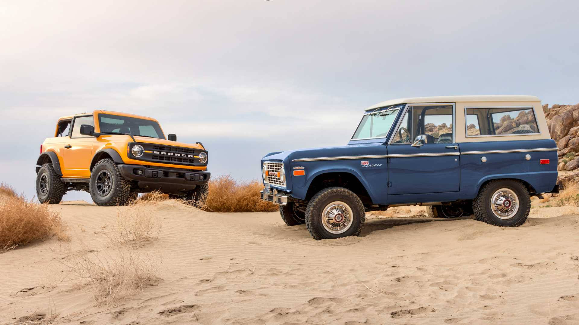 Ford Bronco 桌布 壁紙 — 兩輛復古Bronco，一輛橙色一輛藍色，停放在沙丘上，背景是岩石山丘，天空多雲。