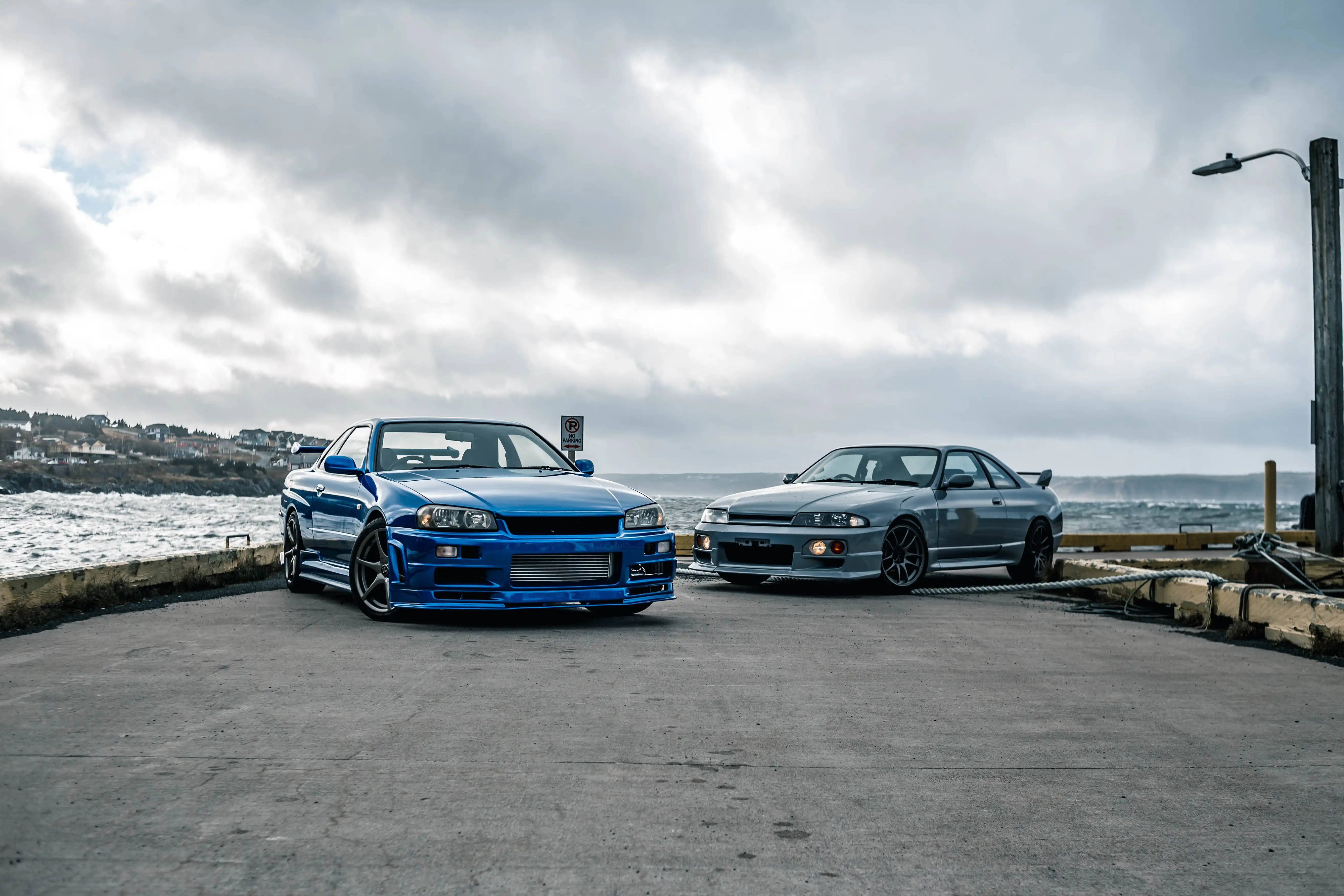 Nissan 桌布 壁紙 — 兩輛改裝的日產Skyline GT-R停泊在海岸碼頭上，天空多雲，海浪在附近拍打。