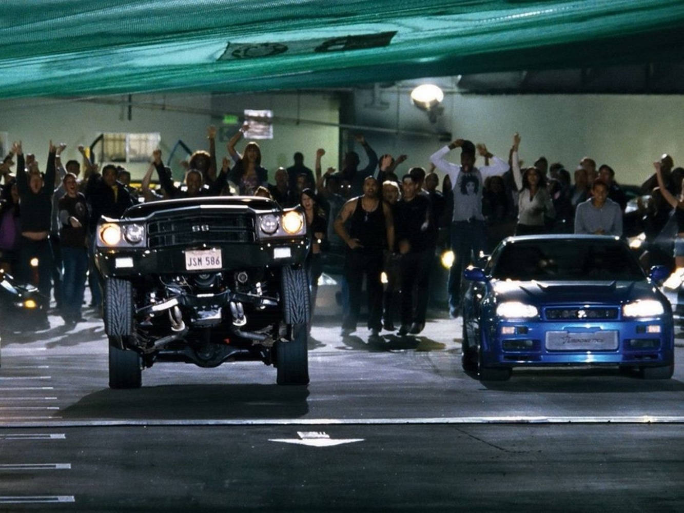 drag race 桌布 壁紙 — 兩輛改裝肌肉車在燈光昏暗的地下車庫中對峙，興奮的人群為他們歡呼。