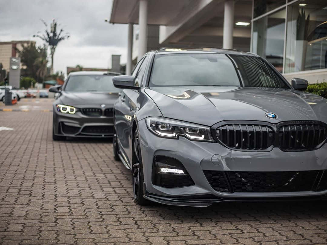 BMW 動圖 桌布 壁紙 — 兩輛時尚灰色BMW停放在圖案磚砌車道上，天空陰沉，背景為現代建築。