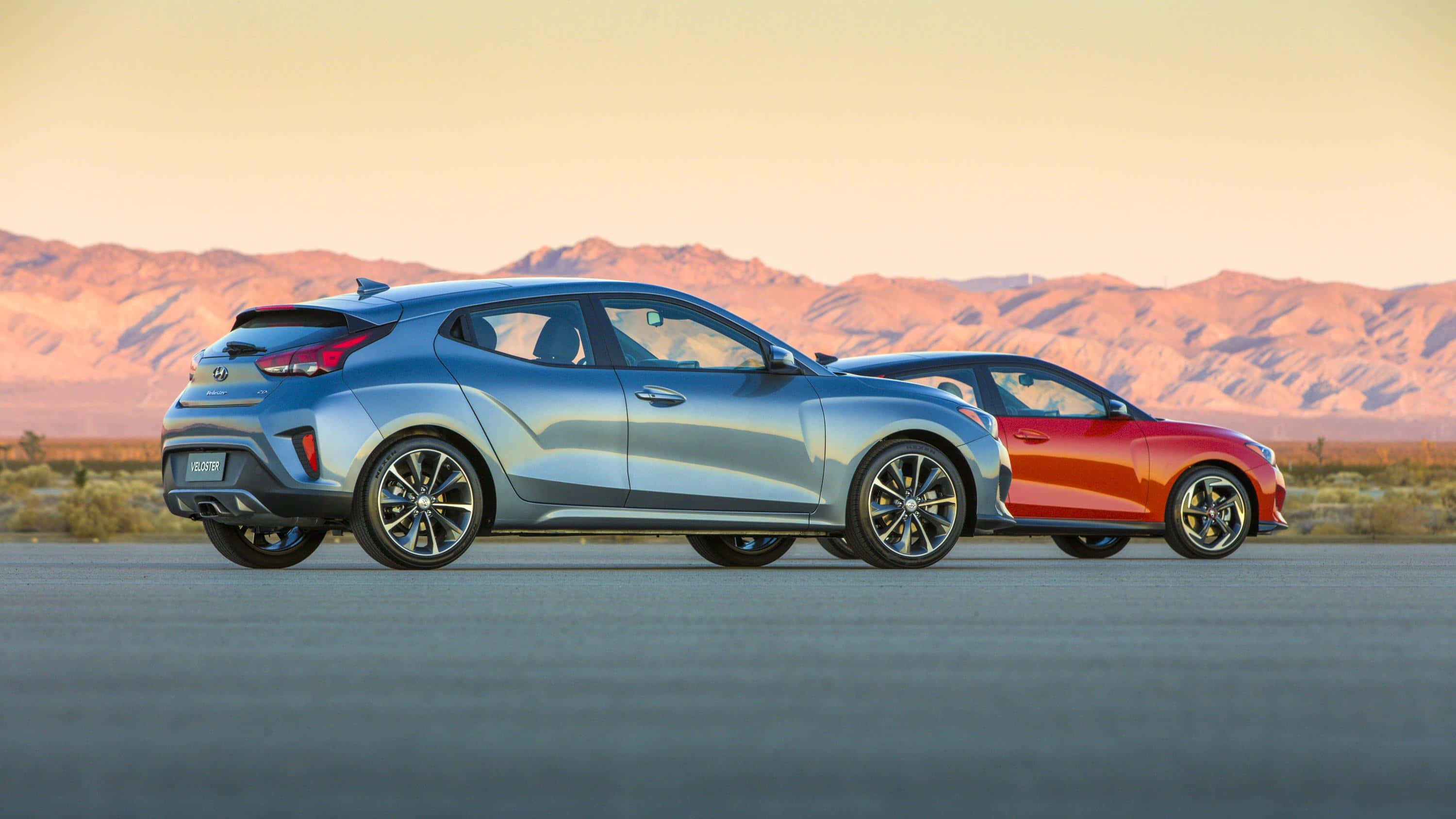 Hyundai Veloster 桌布 壁紙 — 兩輛時尚現代掀背車，銀色和紅色，並排停放在沙漠公路上，背景是日落時分的山脈。