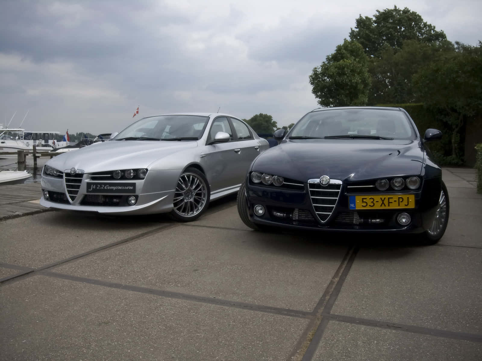 Alfa Romeo 159 桌布 壁紙 — 兩輛時尚的Alfa Romeo轎車，銀色和黑色，並排停放在鋪砌的碼頭上，背景陰天，有船隻和樹木。