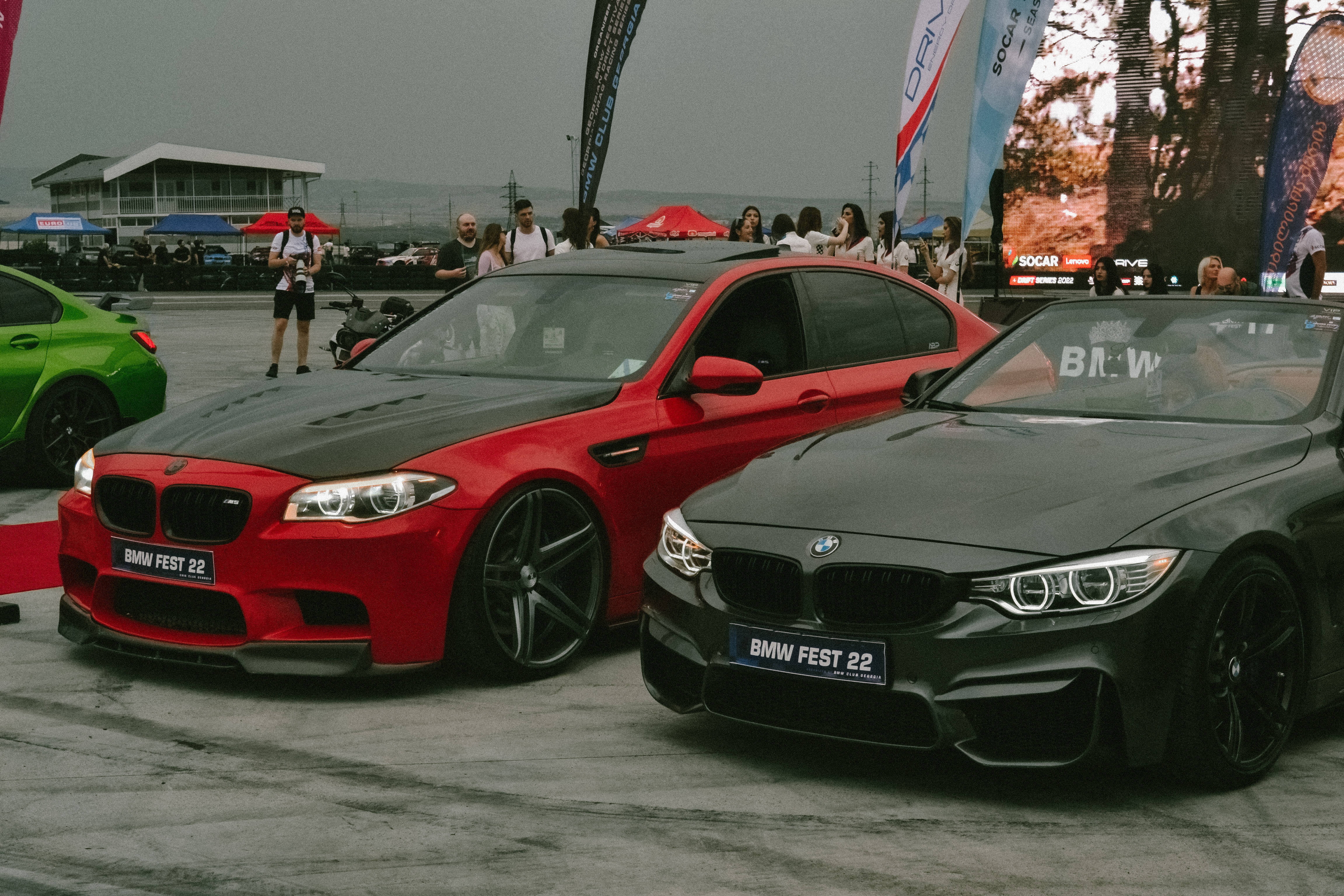 Racing 桌布 壁紙 — 兩輛時尚的BMW紅色和黑色在陰天下的熱鬧車展中閃耀，人群和旗幟環繞。