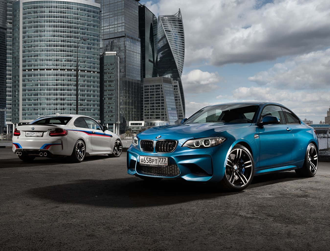 Bmw Tablet 桌布 壁紙 — 兩輛時尚的BMW跑車，一藍一白，停在瀝青路面上，背景是現代玻璃摩天大樓，天空多雲。