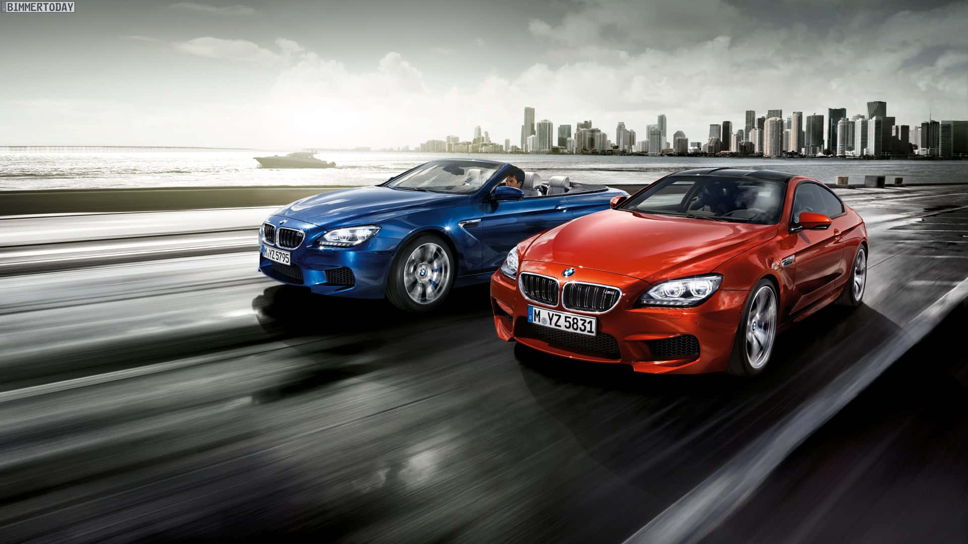 Bmw M Series 桌布 壁紙 — 兩輛時尚的BMW跑車在濕滑的沿海高速公路上奔馳，背景是現代化的城市天際線。