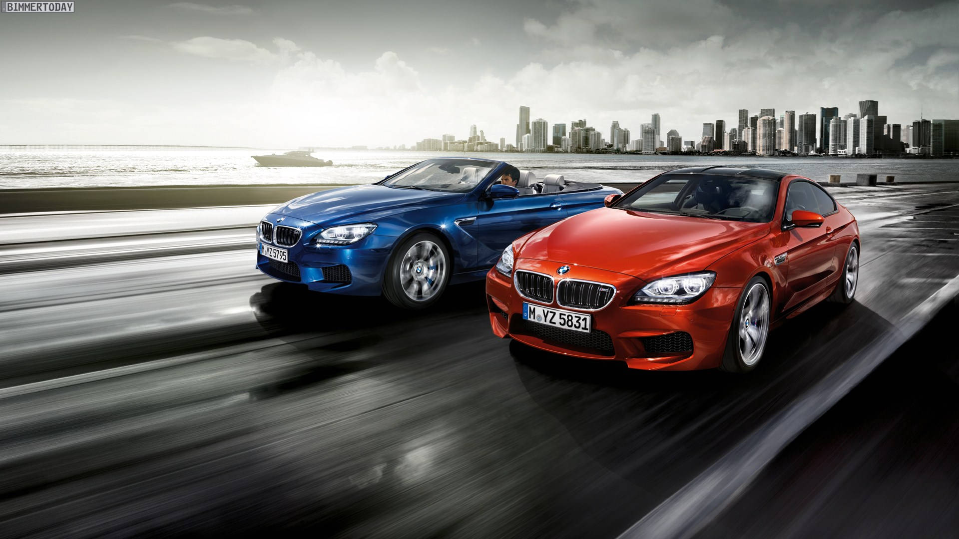 BMW 動圖 桌布 壁紙 — 兩輛時尚的BMW跑車沿著濕漉漉的沿海高速公路奔馳，背景是現代化的城市天際線。