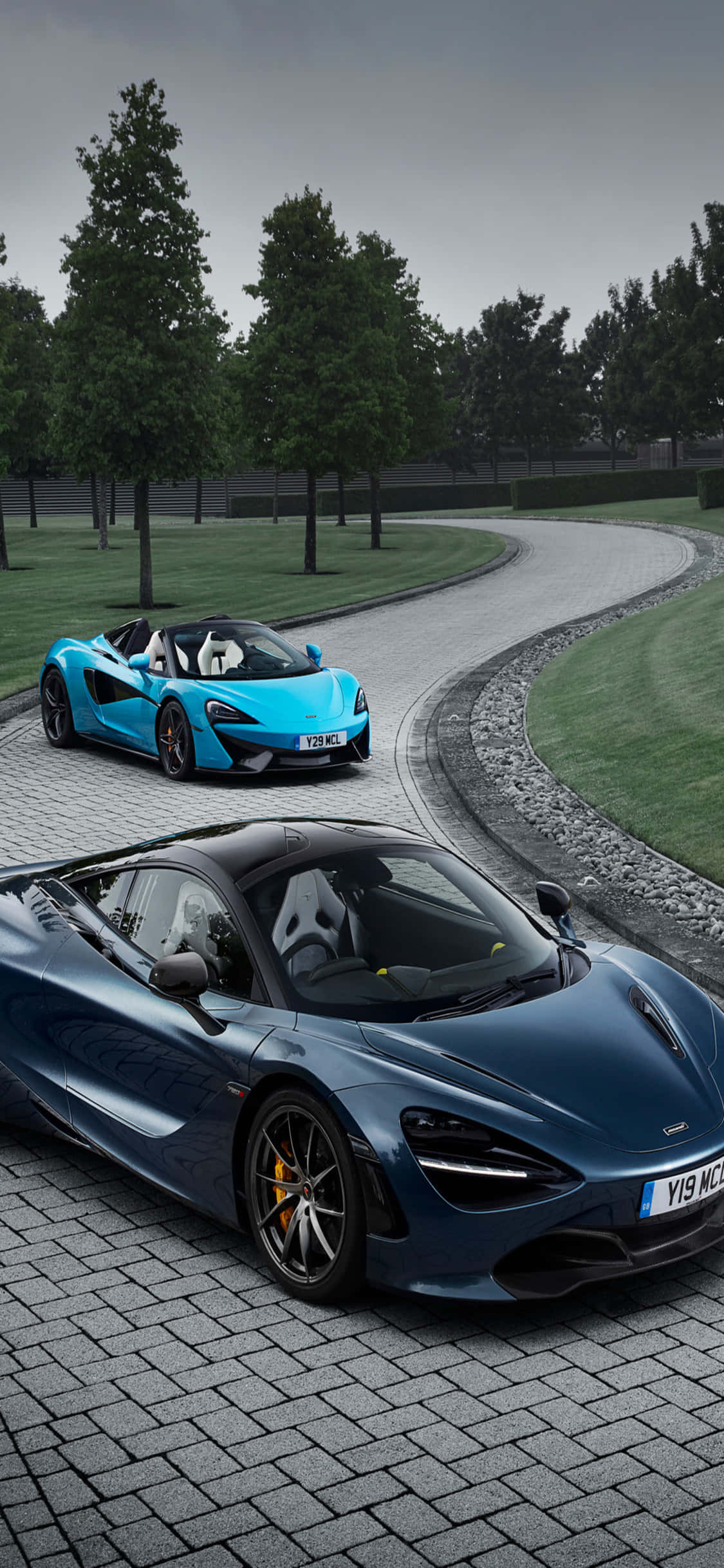 麥拉倫 桌布 壁紙 — 兩輛時尚的McLaren超級跑車，一輛藍色一輛深藍色，停放在彎曲的石頭車道上，周圍綠樹成蔭，天空多雲。
