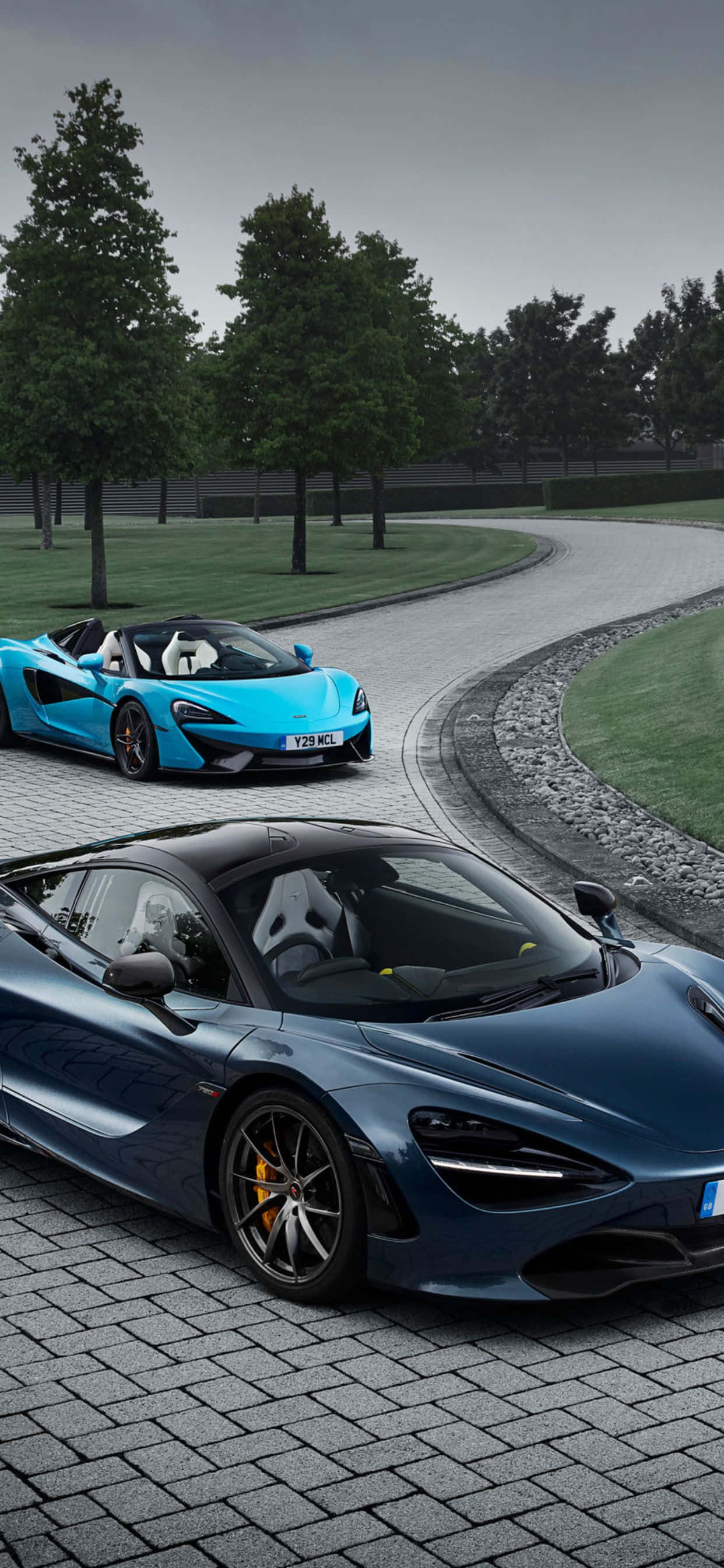 淺藍色 桌布 壁紙 — 兩輛時尚的McLaren跑車，一輛藍色敞篷車和一輛深藍色轎跑車，停放在彎曲的石路上，周圍綠樹成蔭，天空多雲。