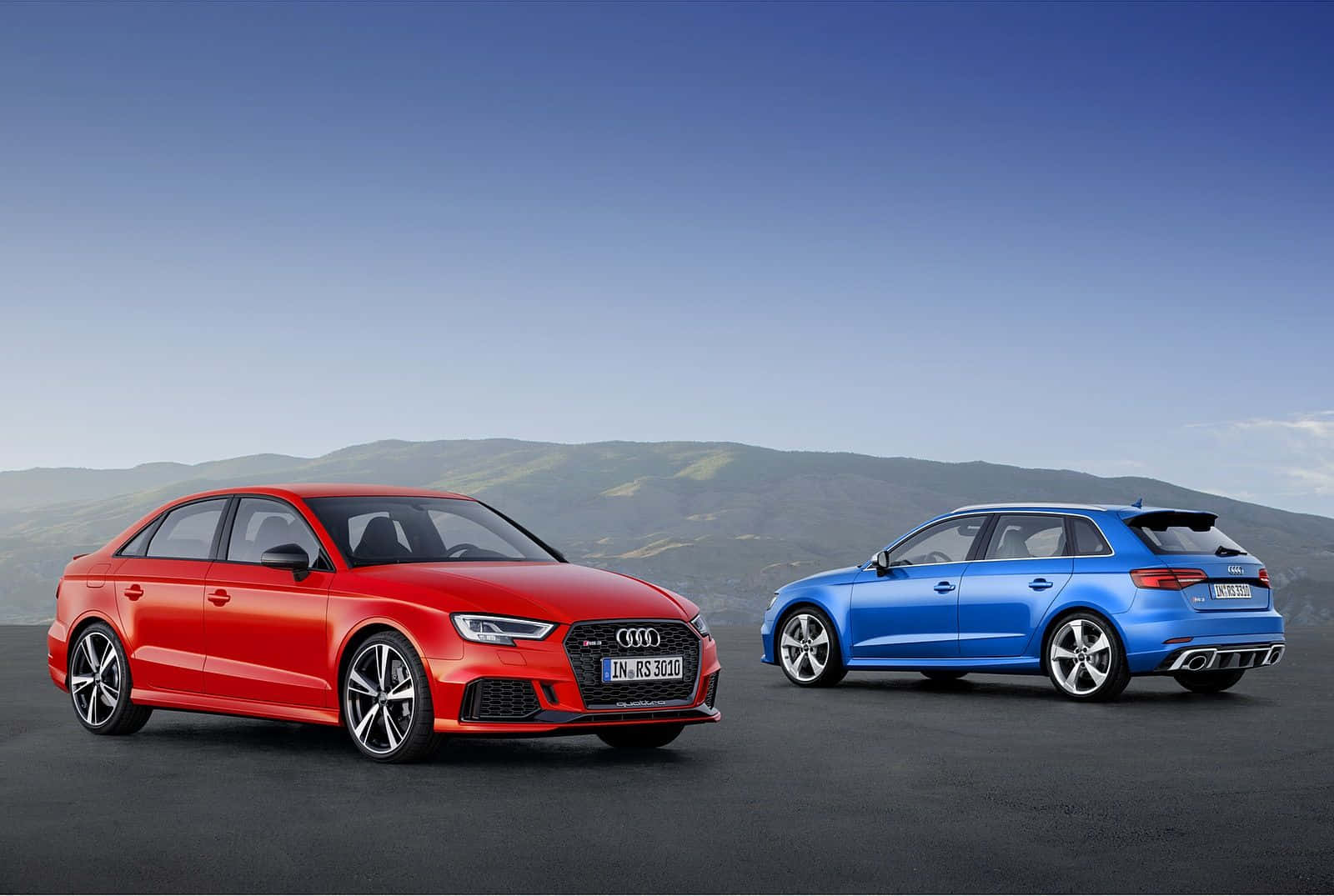Audi Rs3 桌布 壁紙 — 兩輛時尚的奧迪RS3轎車，一紅一藍，停在深色路面上，背景是連綿起伏的綠色山丘和晴朗的藍天。