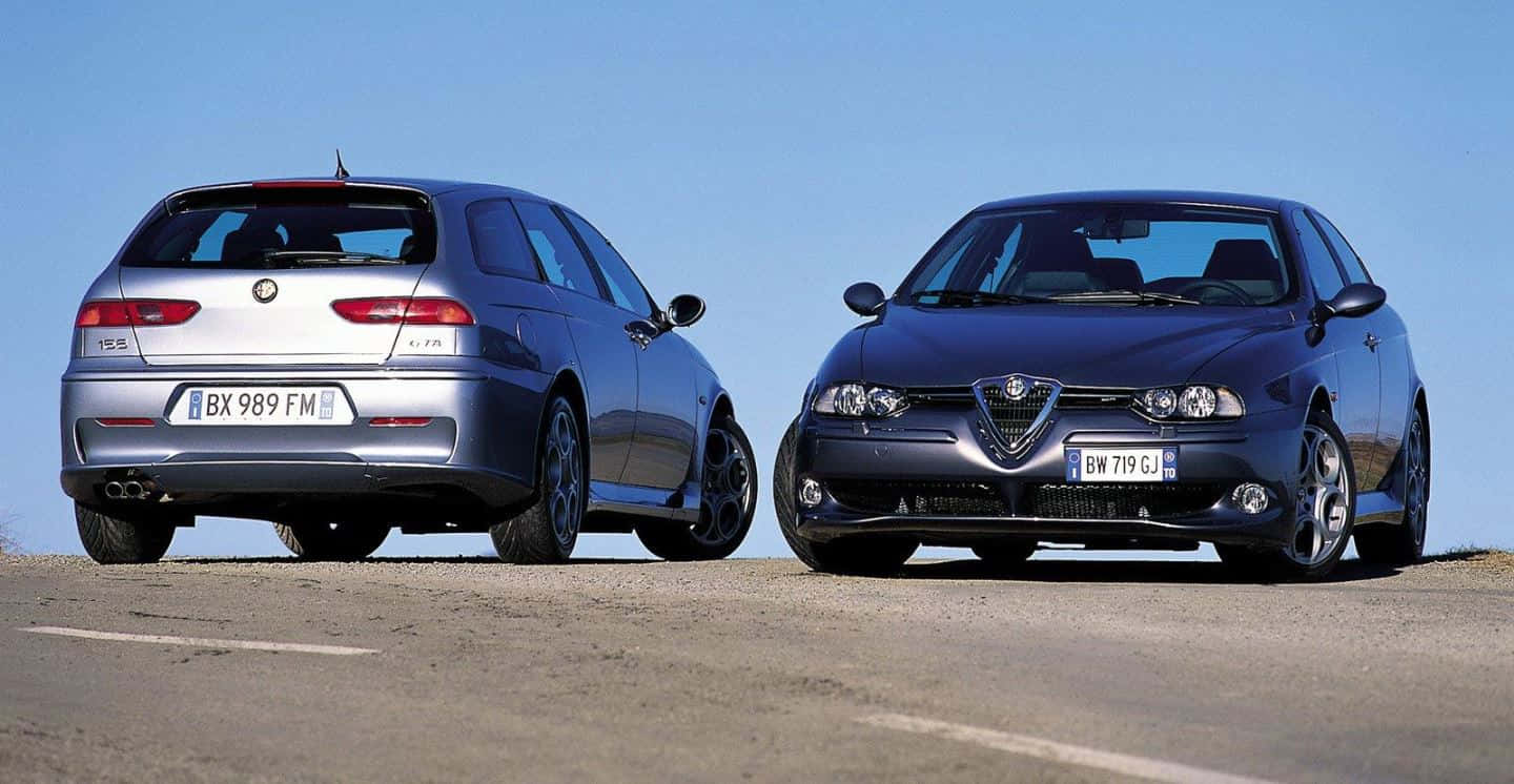 Alfa Romeo 156 桌布 壁紙 — 兩輛時尚的愛快羅密歐汽車（銀色和藍色）並排停放在陽光明媚的道路上，天空清澈湛藍。