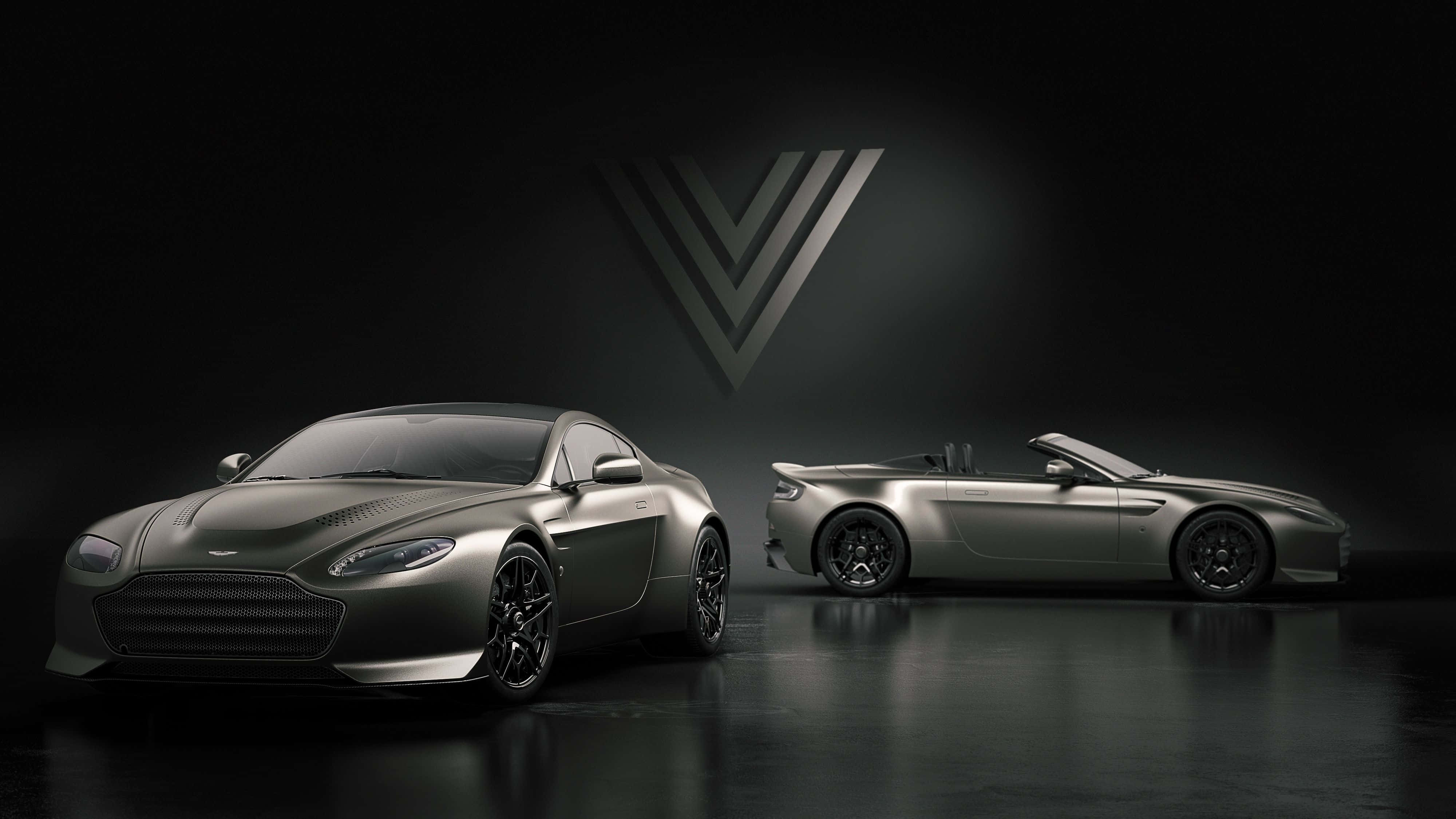 Aston Martin V12 Vantage 桌布 壁紙 — 兩輛時尚的消光灰色 Aston Martin 跑車在黑暗的攝影棚背景下，於戲劇性的聚光燈下閃耀，並帶有發光的 V 形標誌。