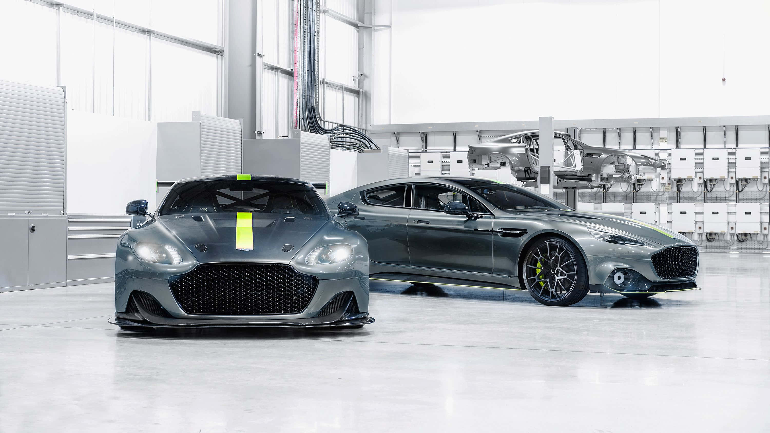 Aston Martin Rapide S 桌布 壁紙 — 兩輛時尚的灰色 Aston Martin 跑車帶有螢光黃色點綴，在極簡工廠環境的明亮燈光下閃閃發光。