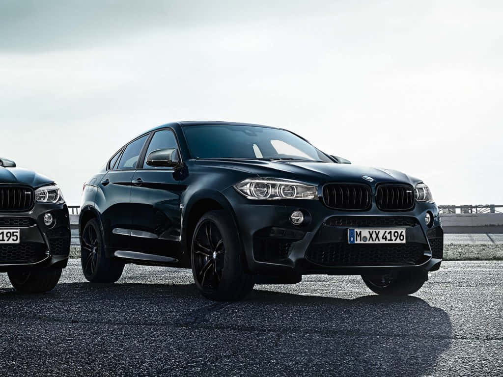 BMW X6 M 桌布 壁紙 — 兩輛時尚的黑色BMW SUV停泊在瀝青路面上，天空多雲，帶有戲劇性的陰影。