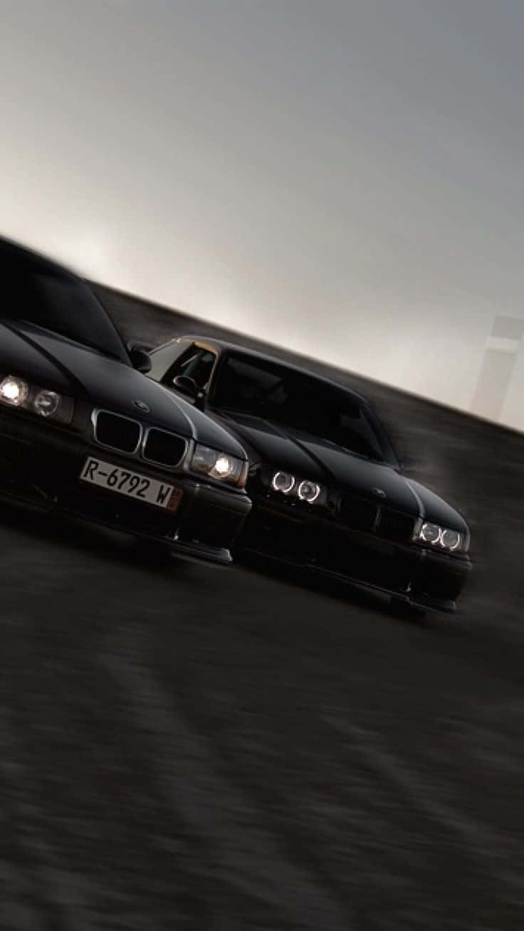 BMW 動圖 桌布 壁紙 — 兩輛時尚的黑色BMW在朦朧的暮色天空下，準備在模糊的瀝青賽道上進行高速對決。