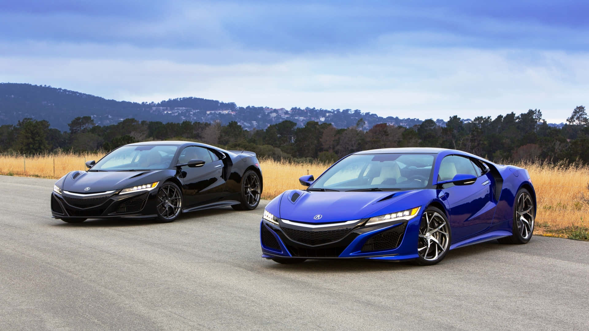 Acura Nsx 桌布 壁紙 — 兩輛時尚跑車，黑色和藍色，停放在一個有山丘和樹木的道路上，天空多雲。