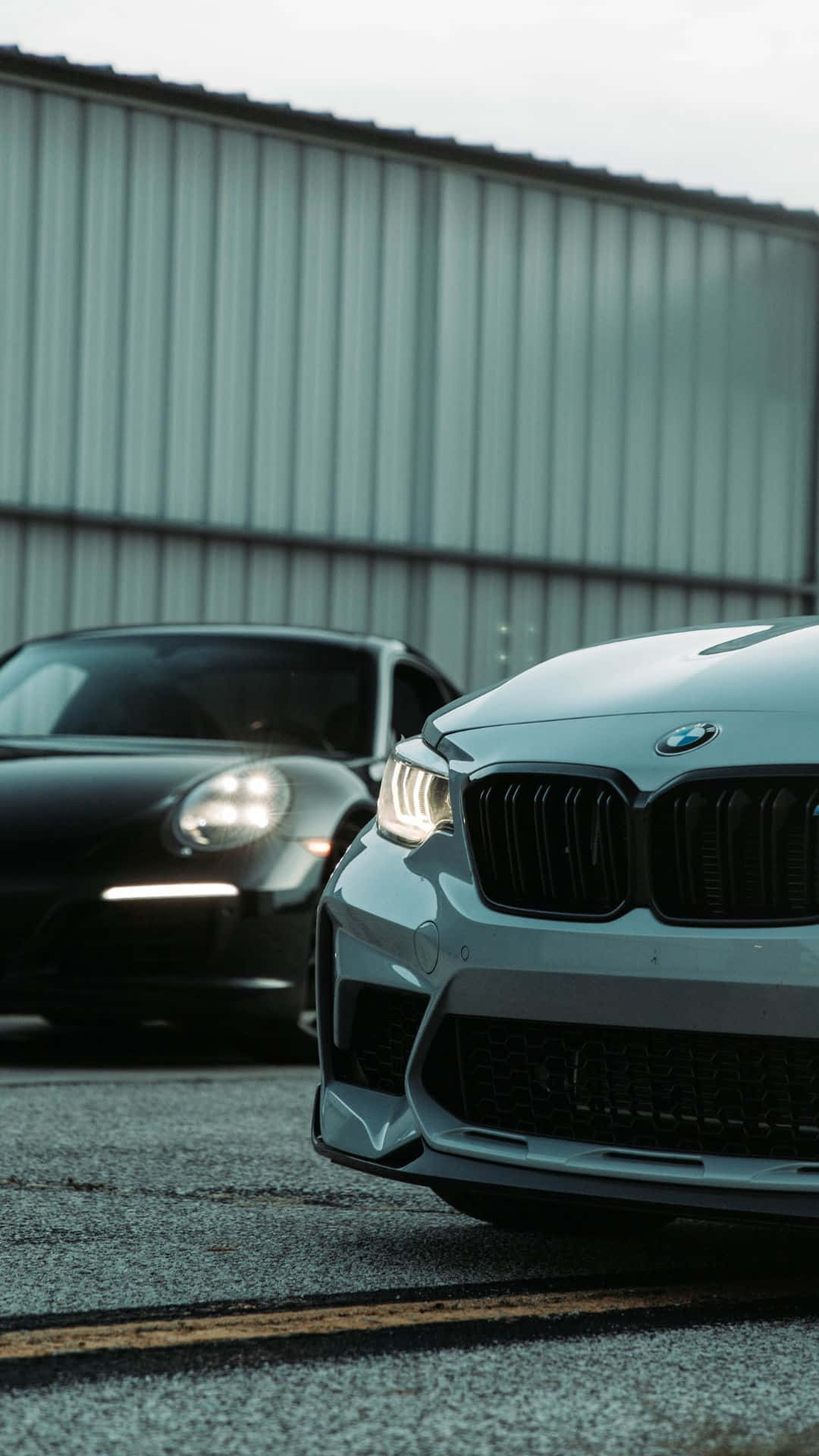 Android Bmw 桌布 壁紙 — 兩輛時尚跑車停在瀝青路上，背景是波紋金屬建築，天空多雲。
