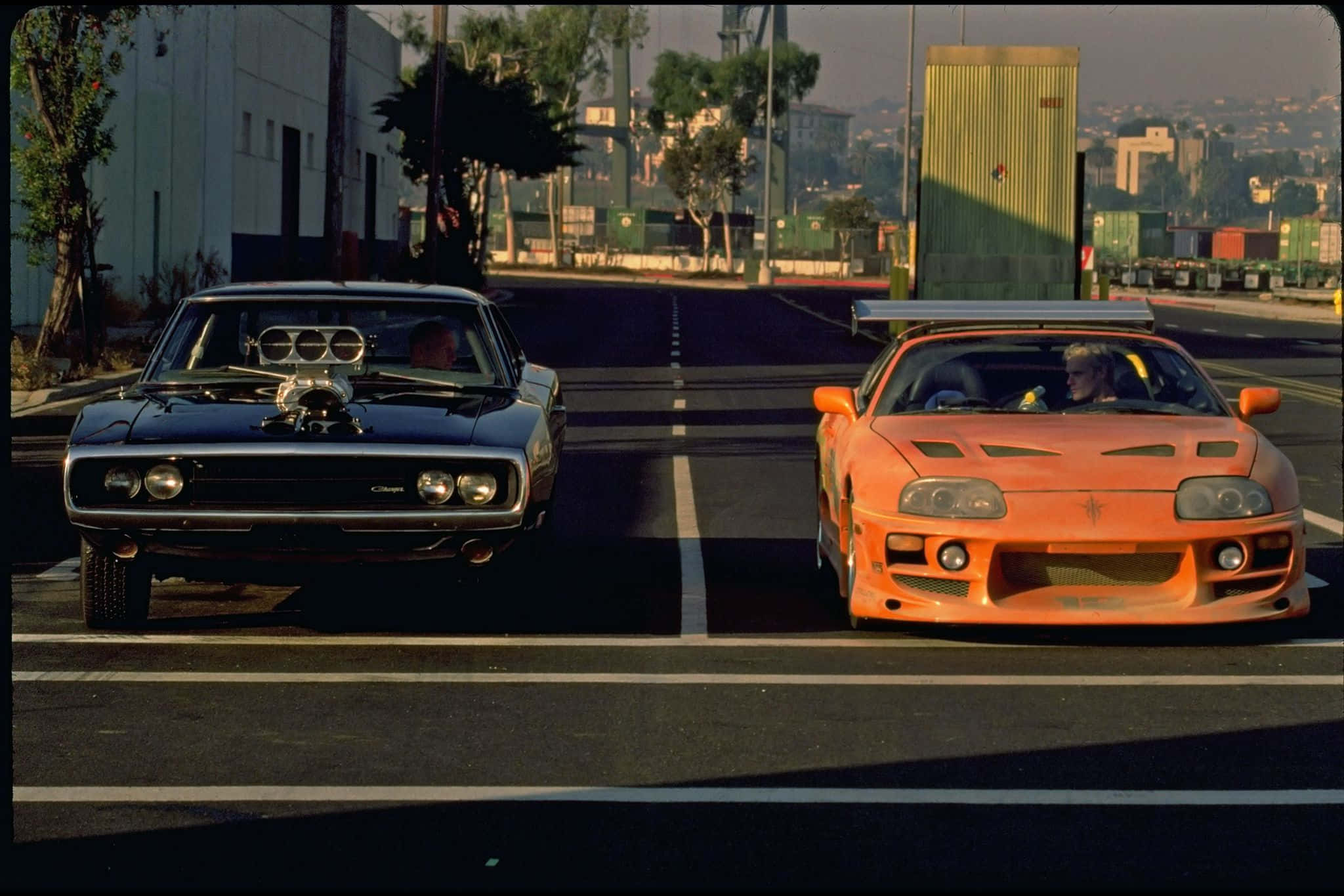 Fast And Furious 1 桌布 壁紙 — 兩輛標誌性的肌肉車在空蕩的高速公路上對峙，背景是工業建築和遠處的城市天際線，沐浴在溫暖的金色光芒中。