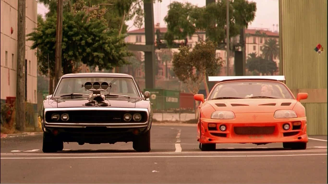 Toyota Supra Mk4 桌布 壁紙 — 兩輛標誌性的肌肉車在陽光明媚、兩旁種滿樹木和建築物的城市街道上對峙。