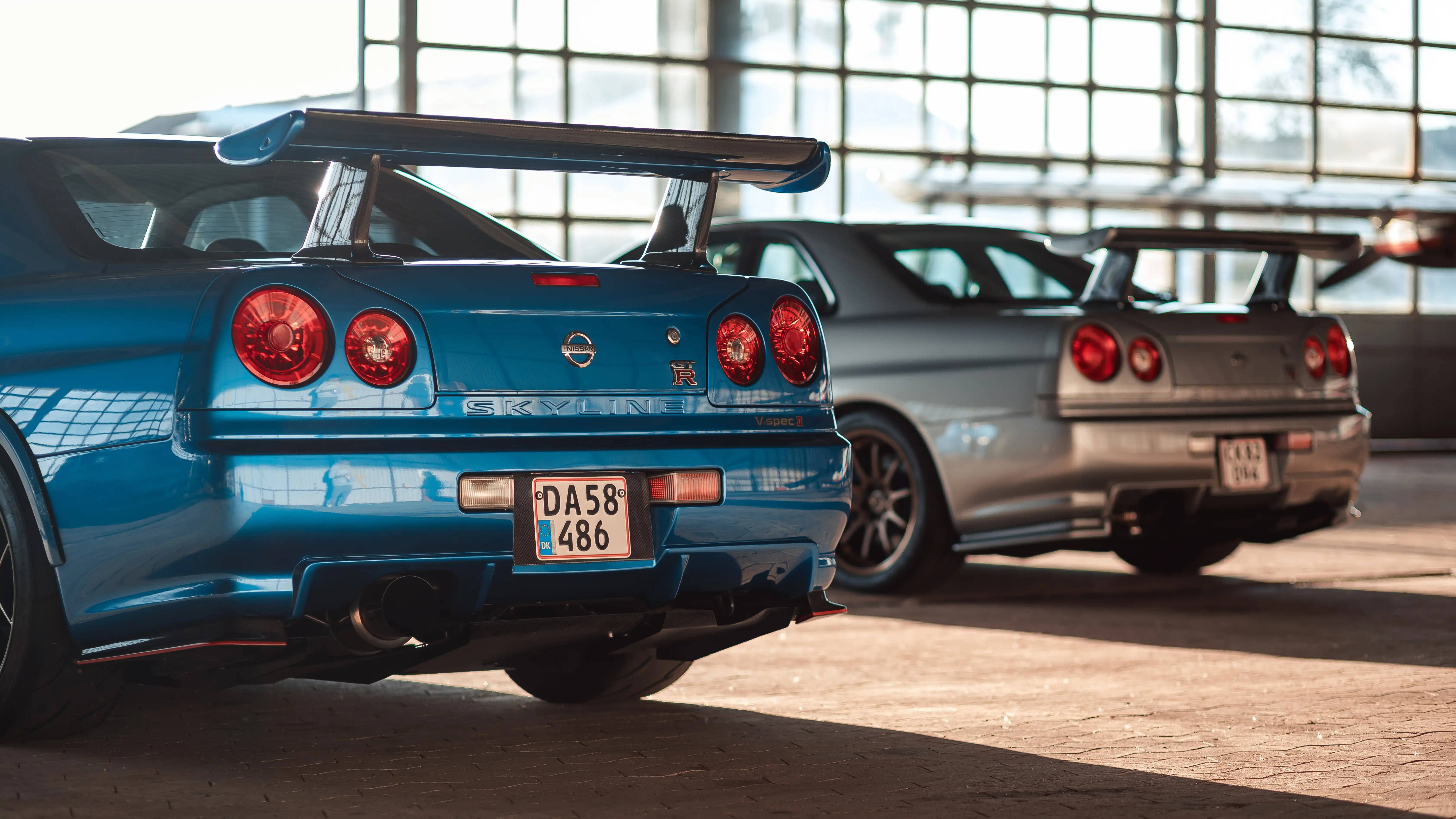 Nissan 桌布 壁紙 — 兩輛標誌性的藍色和銀色日產Skyline GT-R停放在室內，陽光透過大窗戶照射進來。