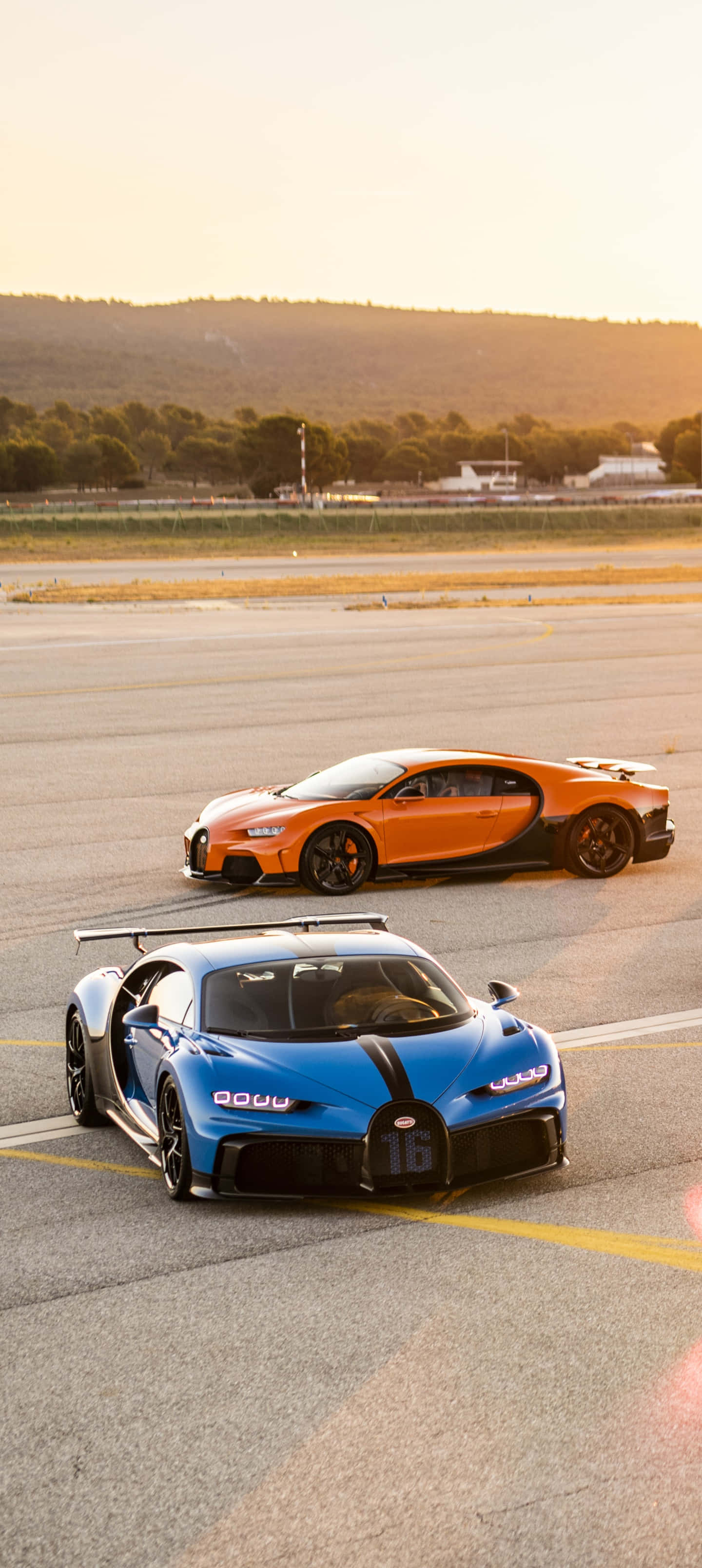 Bugatti Phone 桌布 壁紙 — 兩輛橙色和藍色的超級跑車在黃金時段停泊在跑道上，背景有山丘。