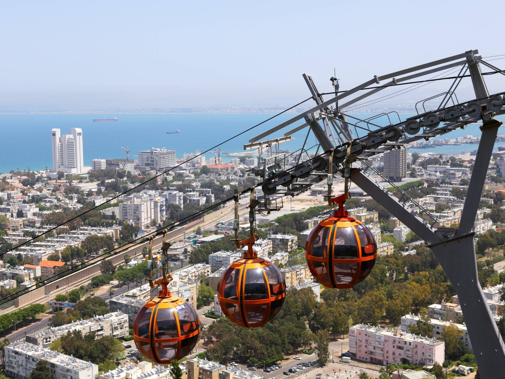 Israel 桌布 壁紙 — 兩輛橙色纜車在陽光明媚的海岸城市景觀上空滑翔，下方是藍色的大海和白色的建築物。