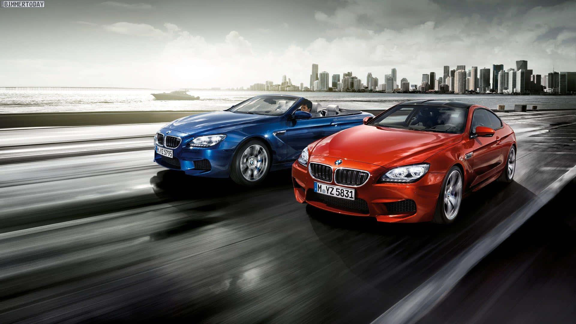 1920x1080 M Series 桌布 壁紙 — 兩輛流線型BMW跑車沿著濕漉漉的海岸高速公路疾馳，背景是城市天際線和海洋。