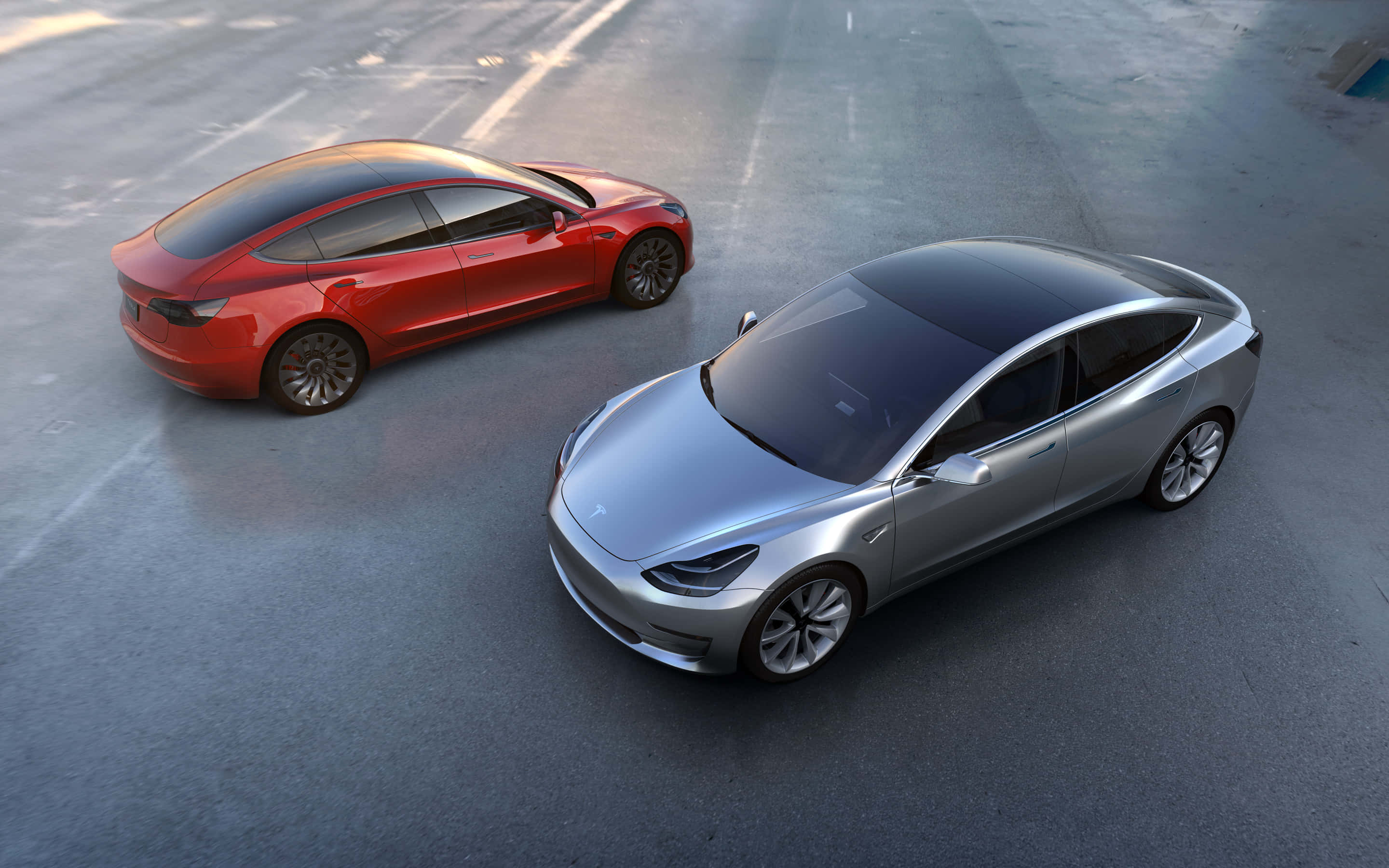 Tesla Model 3 桌布 壁紙 — 兩輛流線型Tesla Model 3轎車，一紅一銀，停在混凝土停車場，柔和日光下。