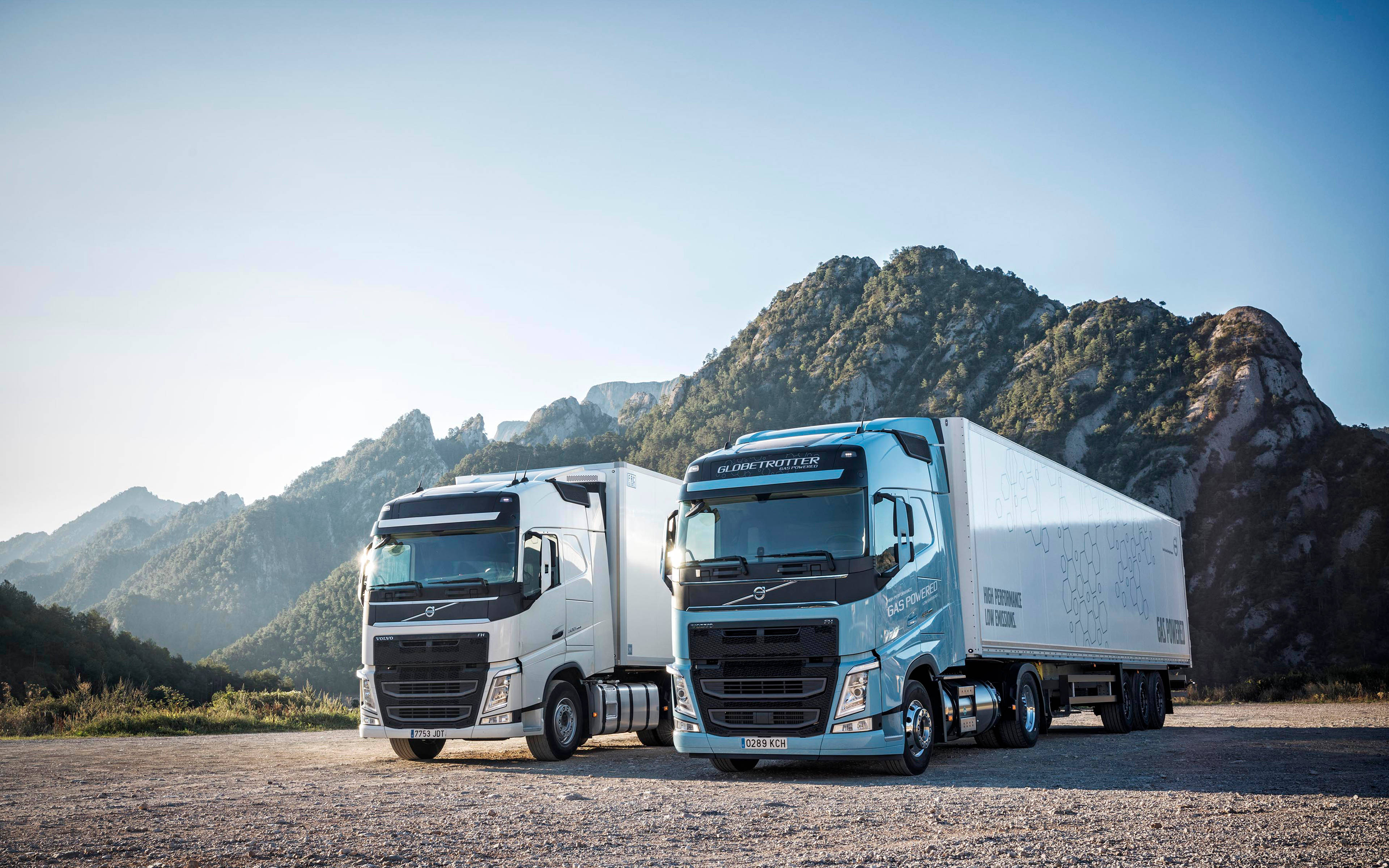 Trucks 桌布 壁紙 — 兩輛現代富豪貨車，一白一藍，停泊在碎石上，背景是崎嶇的山脈，天空清澈湛藍。
