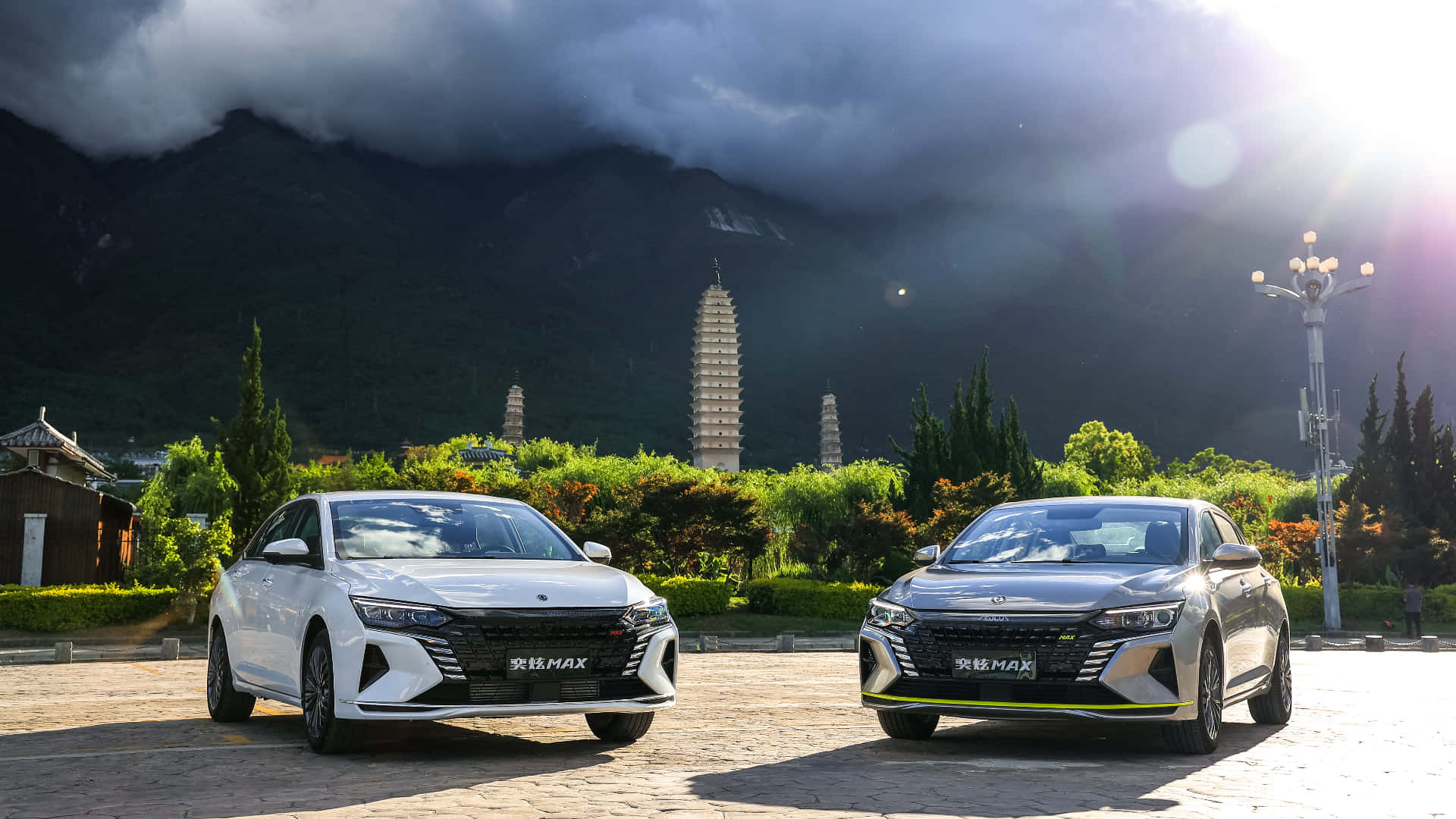 Dongfeng 桌布 壁紙 — 兩輛現代轎車停放在石頭廣場上，背景是霧氣繚繞的山脈和古老的寶塔，天空雲層變幻，氣氛戲劇化。