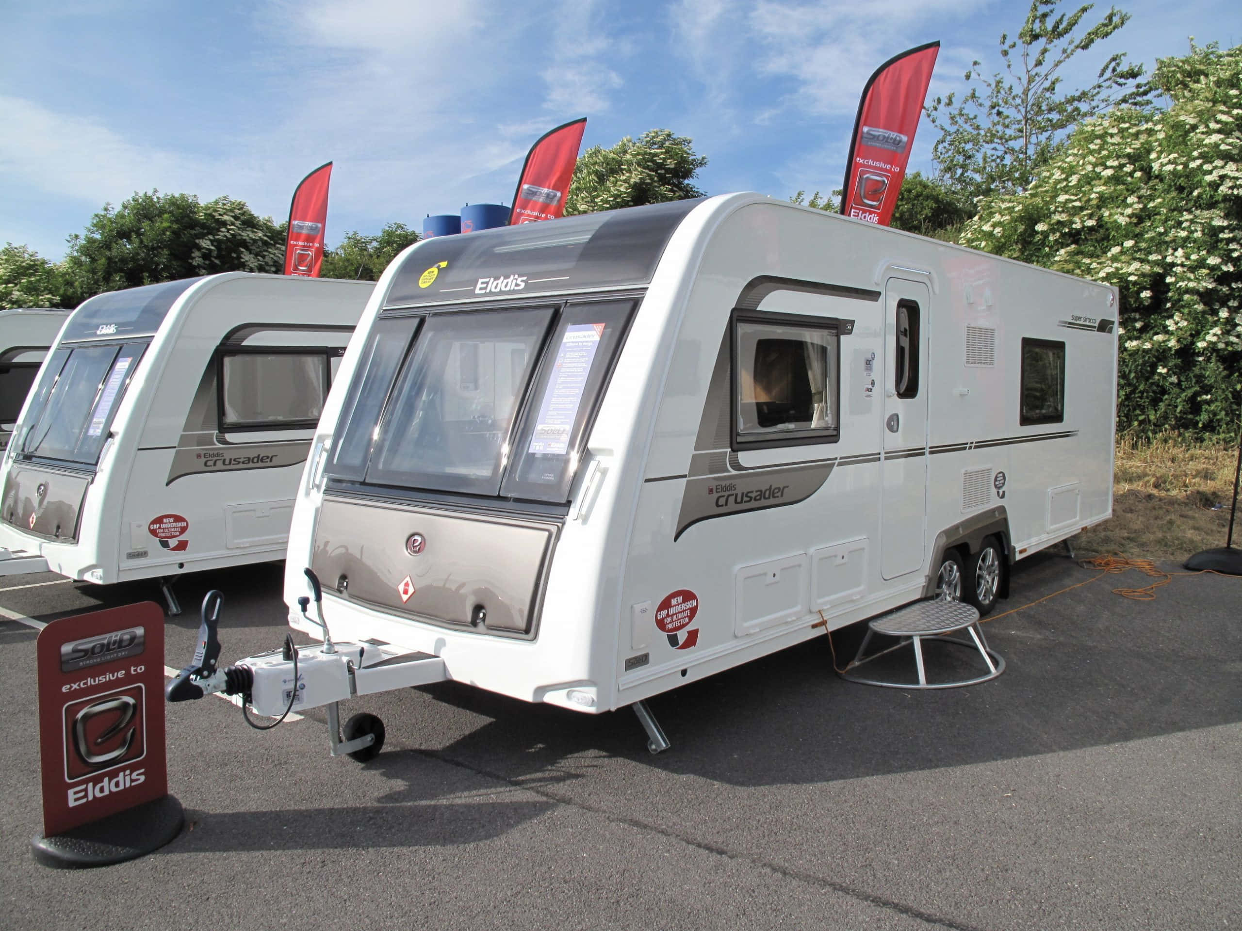 Caravan 桌布 壁紙 — 兩輛白色Elddis Crusader露營車停在瀝青路上，背景是明亮的藍天、紅旗和綠色灌木。