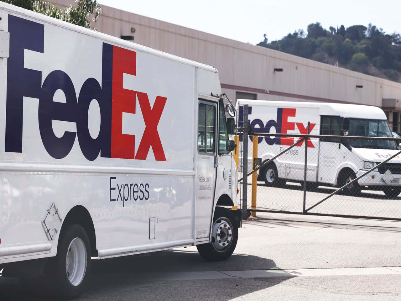 聯邦快遞 桌布 壁紙 — 兩輛白色FedEx Express送貨車停在倉庫外，背景有山丘，光線明亮。
