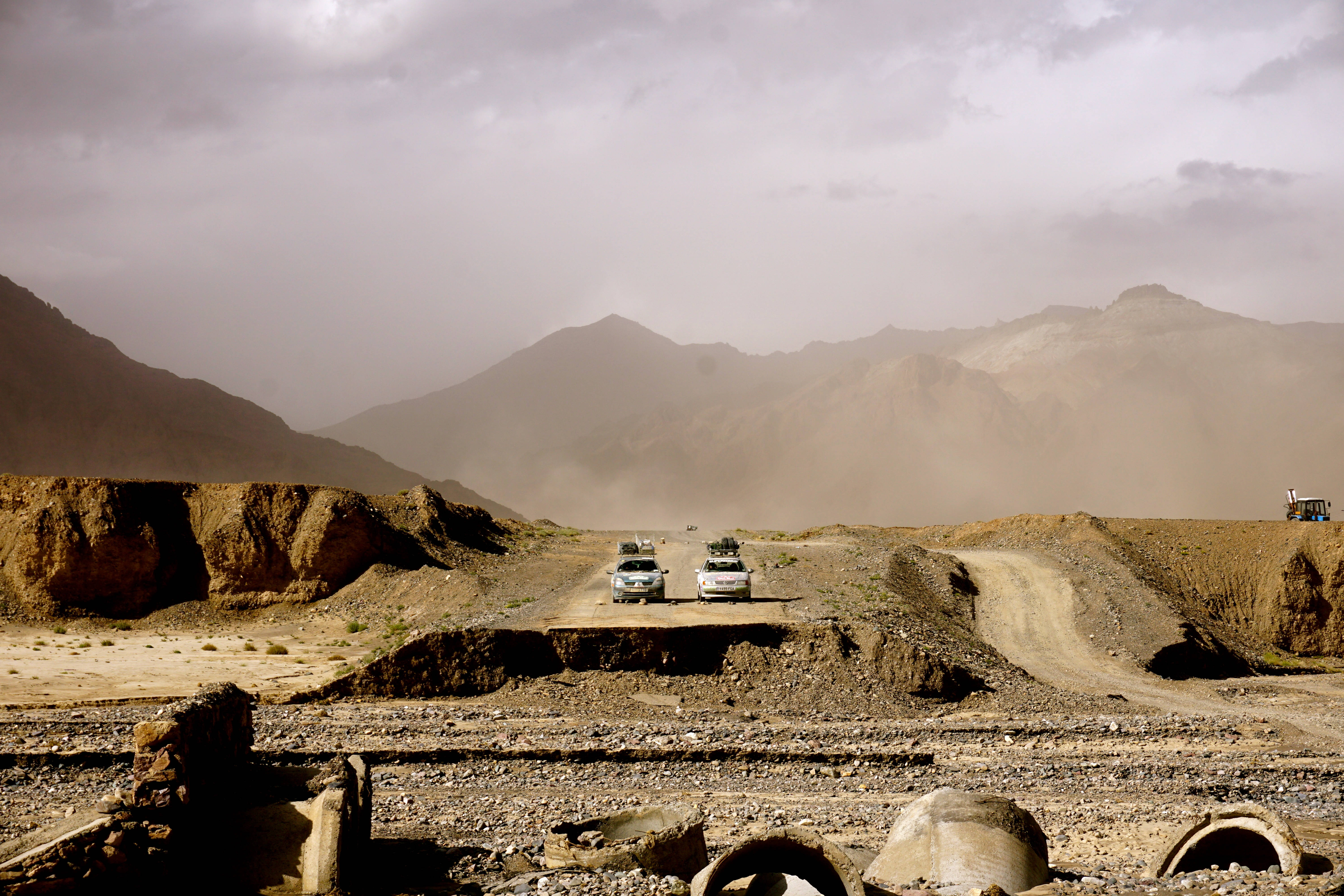 rally 桌布 壁紙 — 兩輛白色SUV沿著塵土飛揚的山路行駛，穿過一片荒蕪的沙漠景觀，天空朦朧。