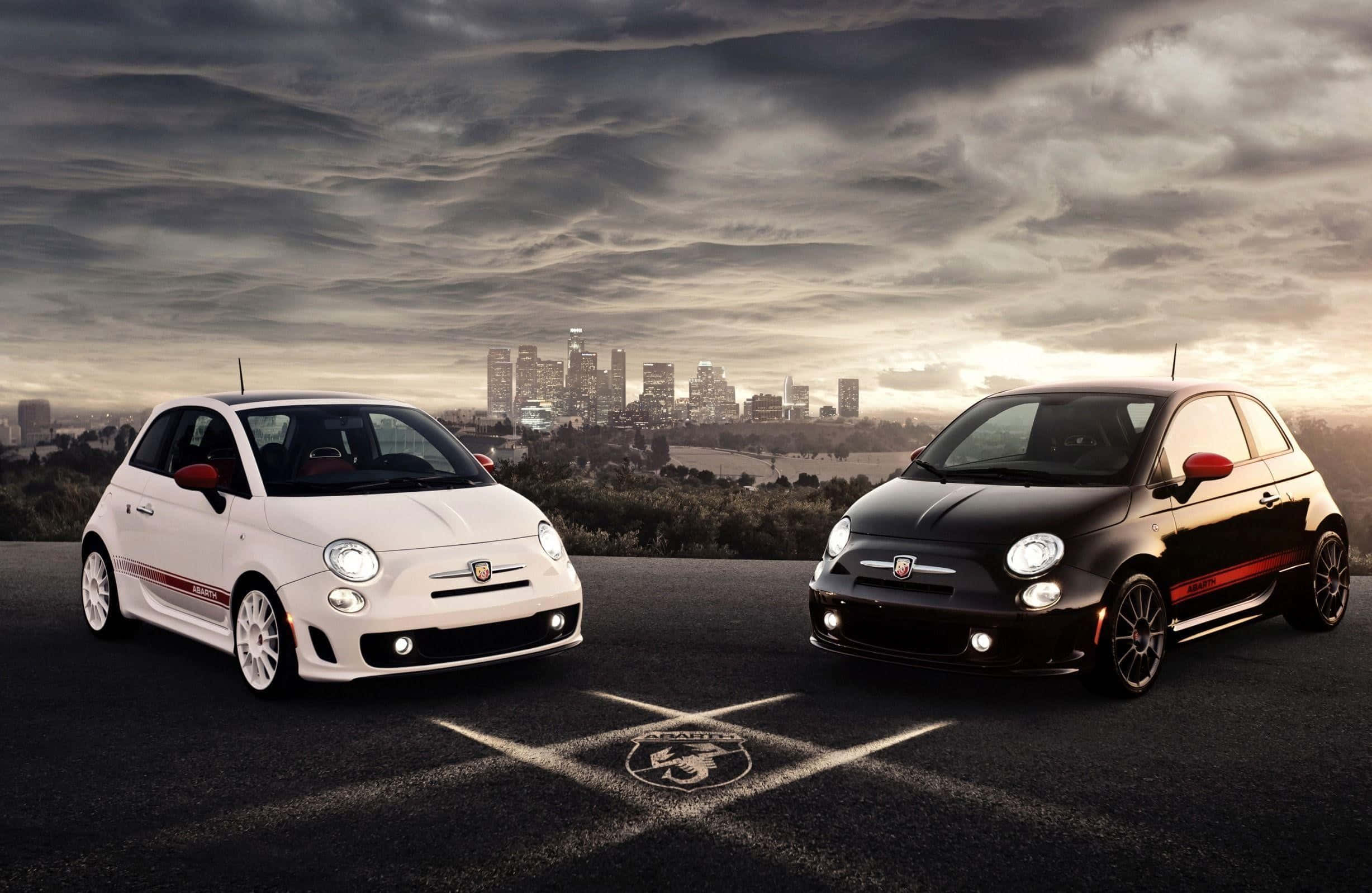 Abarth 桌布 壁紙 — 兩輛白色和黑色帶紅色點綴的Fiat 500汽車停在瀝青路上，背景是戲劇性多雲天空下的城市天際線。