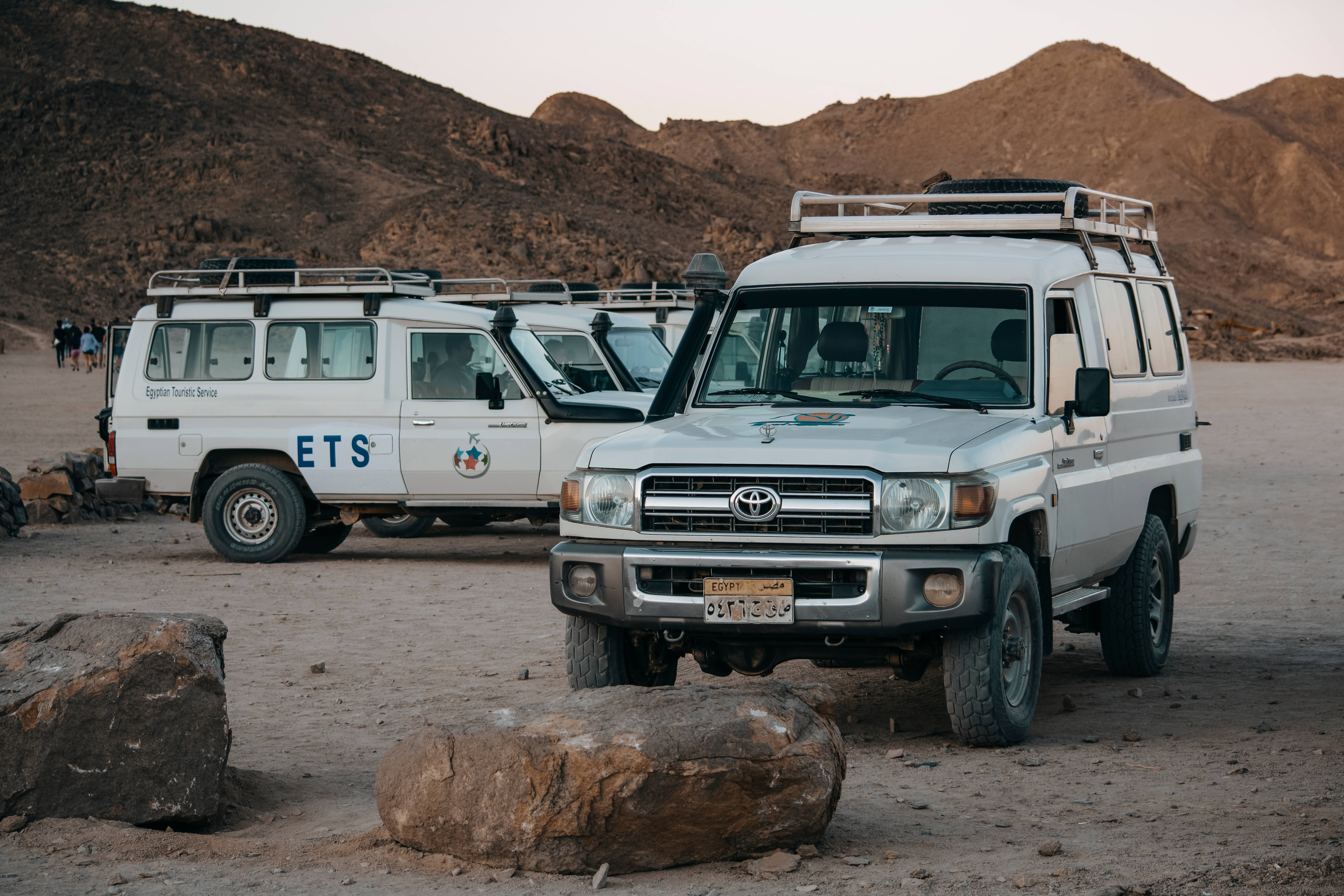Sahara 桌布 壁紙 — 兩輛白色獵遊車停泊在岩石沙漠地形上，背景是黃昏時分崎嶇的山脈。