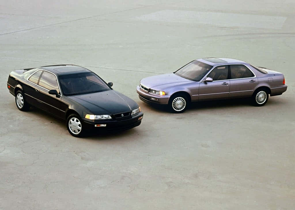 Acura Legend 桌布 壁紙 — 兩輛經典1990年代轎車，黑色雙門跑車和銀色轎車，停在明亮的日光下裂開的混凝土上。
