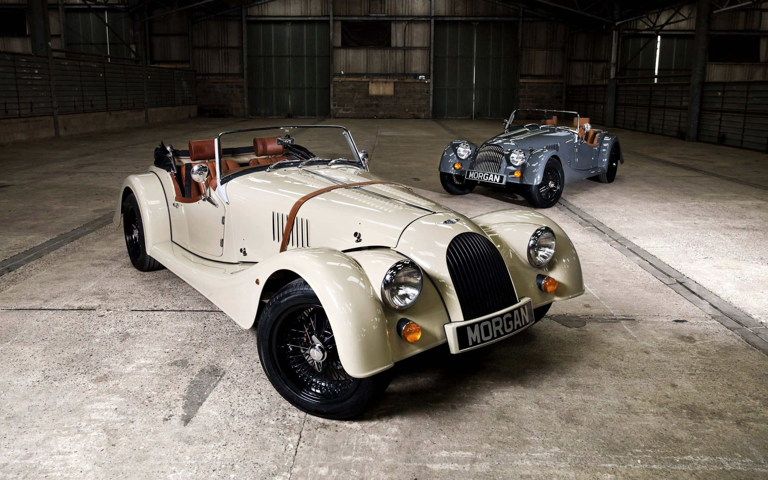 Morgan 桌布 壁紙 — 兩輛經典摩根跑車在寬敞倉庫的工業照明下閃閃發光，倉庫內有混凝土地板和金屬橫樑。