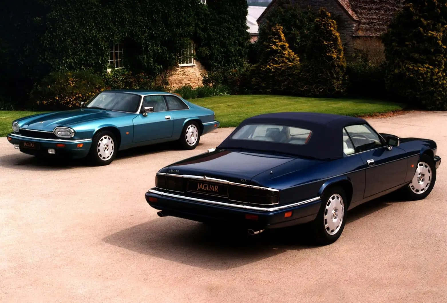 Jaguar Xjs 桌布 壁紙 — 兩輛經典積架汽車，一輛青綠色雙門轎跑車和一輛深藍色敞篷車，停放在石屋前的碎石車道上，周圍綠意盎然。