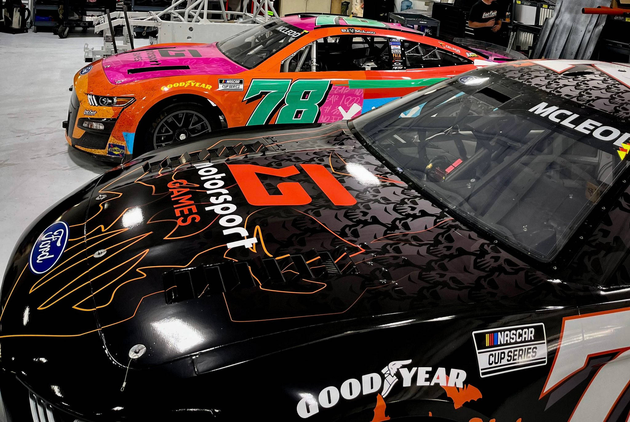 黑色 桌布 壁紙 — 兩輛色彩鮮豔的NASCAR賽車，帶有醒目圖案和贊助商標誌，停放在明亮燈光下的車庫中。