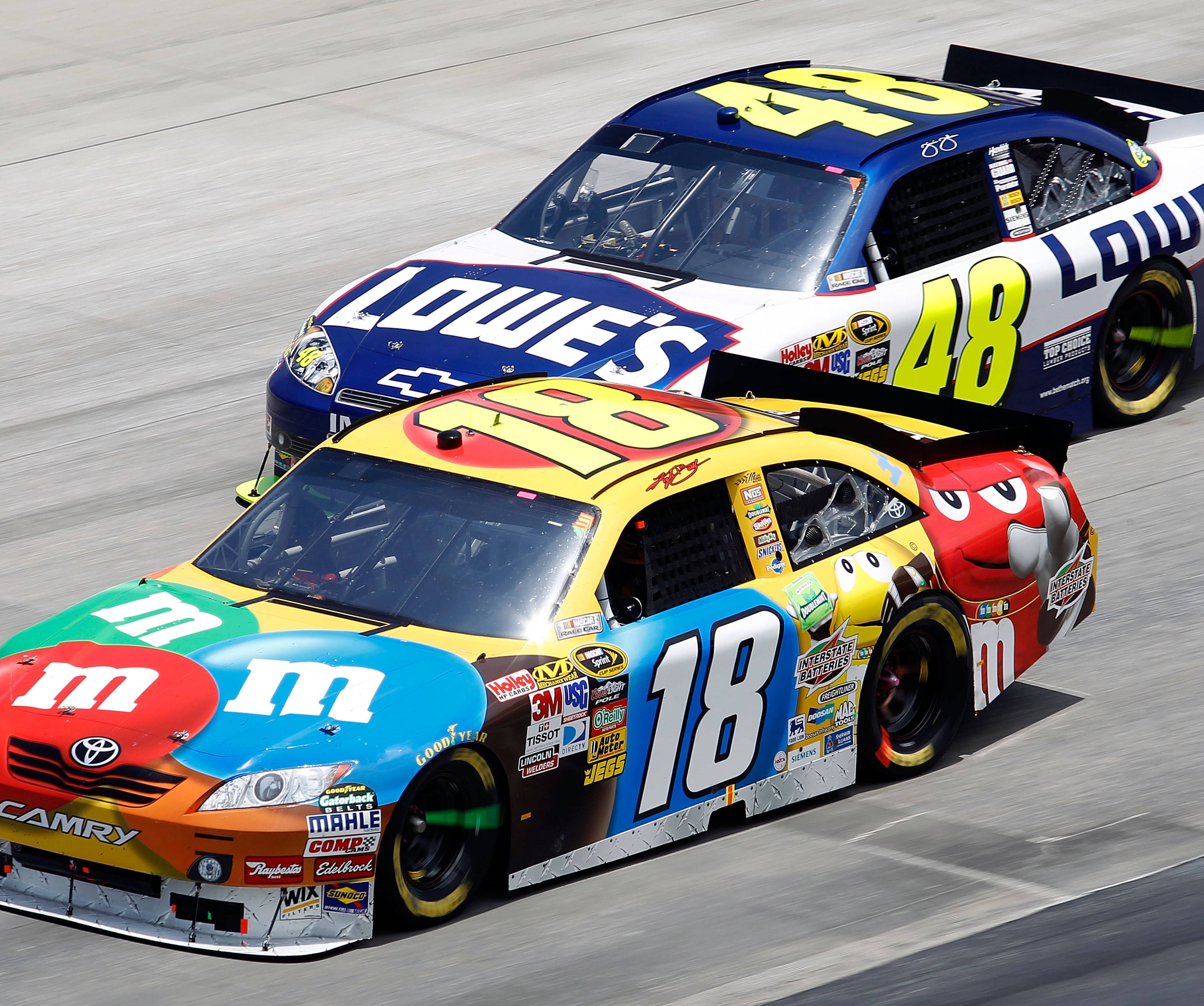Racing 桌布 壁紙 — 兩輛色彩鮮豔的NASCAR賽車在陽光明媚的賽道上並排行駛，一輛飾有M&M's品牌標誌，另一輛帶有Lowe's標誌。