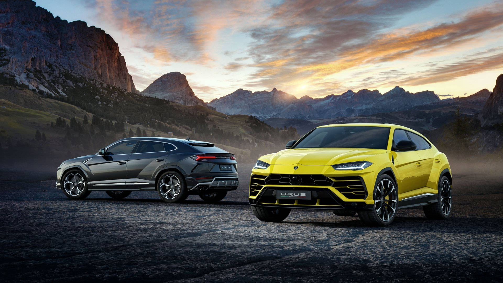 lambo 桌布 壁紙 — 兩輛藍寶堅尼Urus SUV，一輛黃色一輛灰色，停在山路上，夕陽西下，背後有戲劇性的雲彩和岩石山峰。