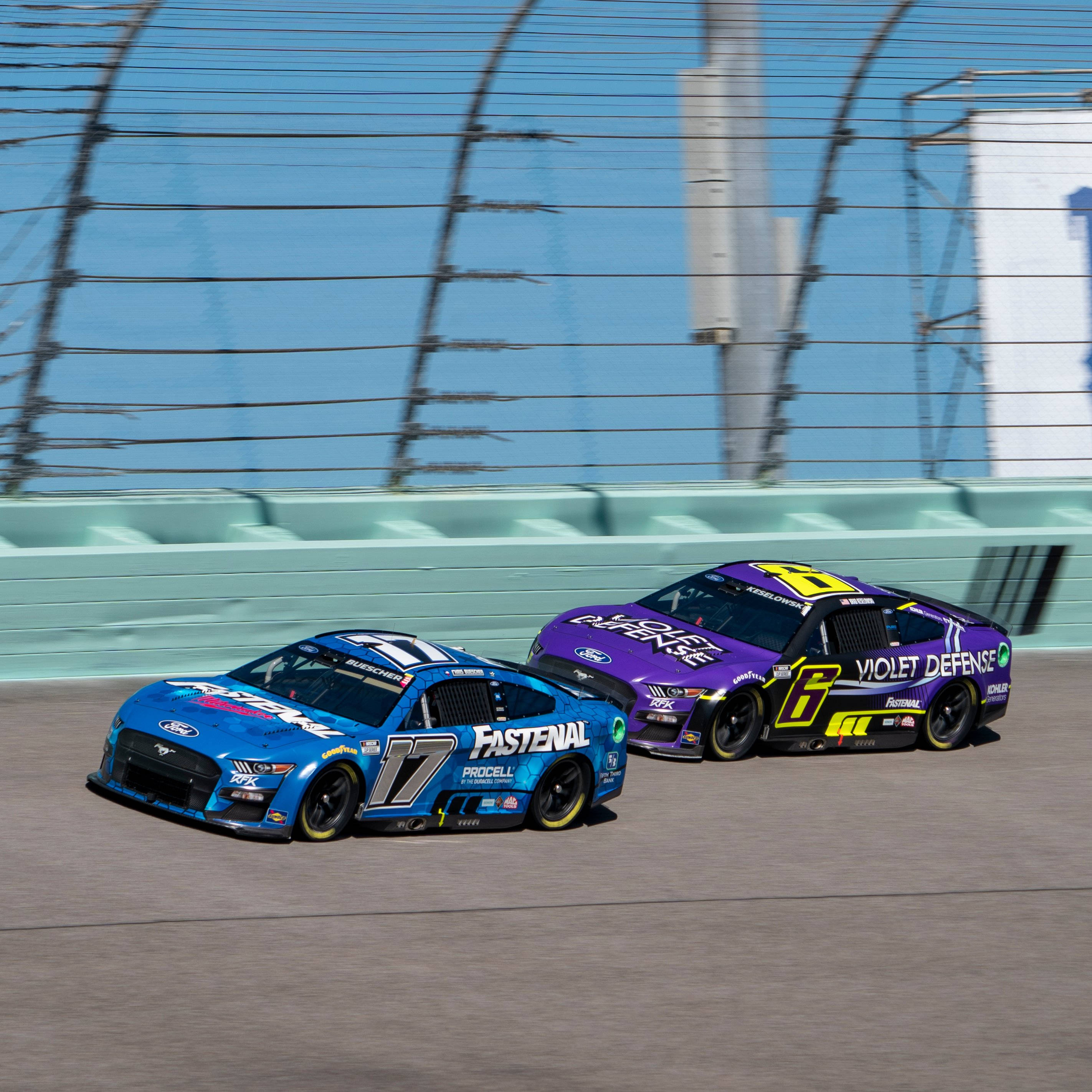 Racing 桌布 壁紙 — 兩輛藍色和紫色的NASCAR賽車在陽光明媚的賽道上並排飛馳，背景是模糊的安全圍欄。