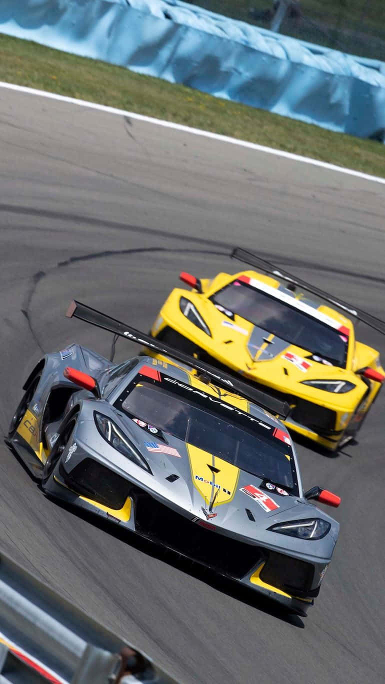race car 桌布 壁紙 — 兩輛賽車在陽光明媚的賽道上激烈角逐，賽道設有藍色安全護欄和模糊的瀝青動感。