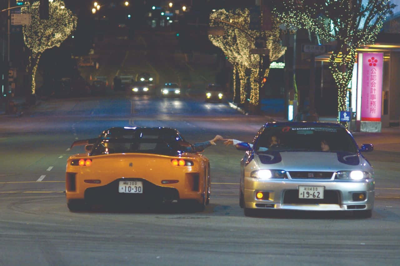 Fast And Furious 桌布 壁紙 — 兩輛跑車在節慶的城市街道上夜間對峙，樹燈閃爍，駕駛互相伸手。