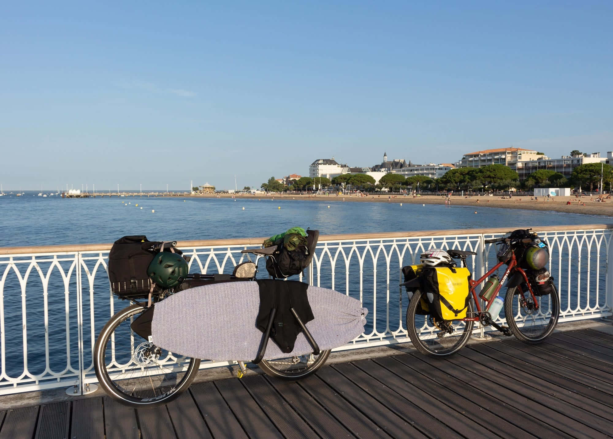 bridge 桌布 壁紙 — 兩輛載滿裝備和衝浪板的旅行自行車停放在木製碼頭上，俯瞰著晴朗天空下平靜的藍色大海。