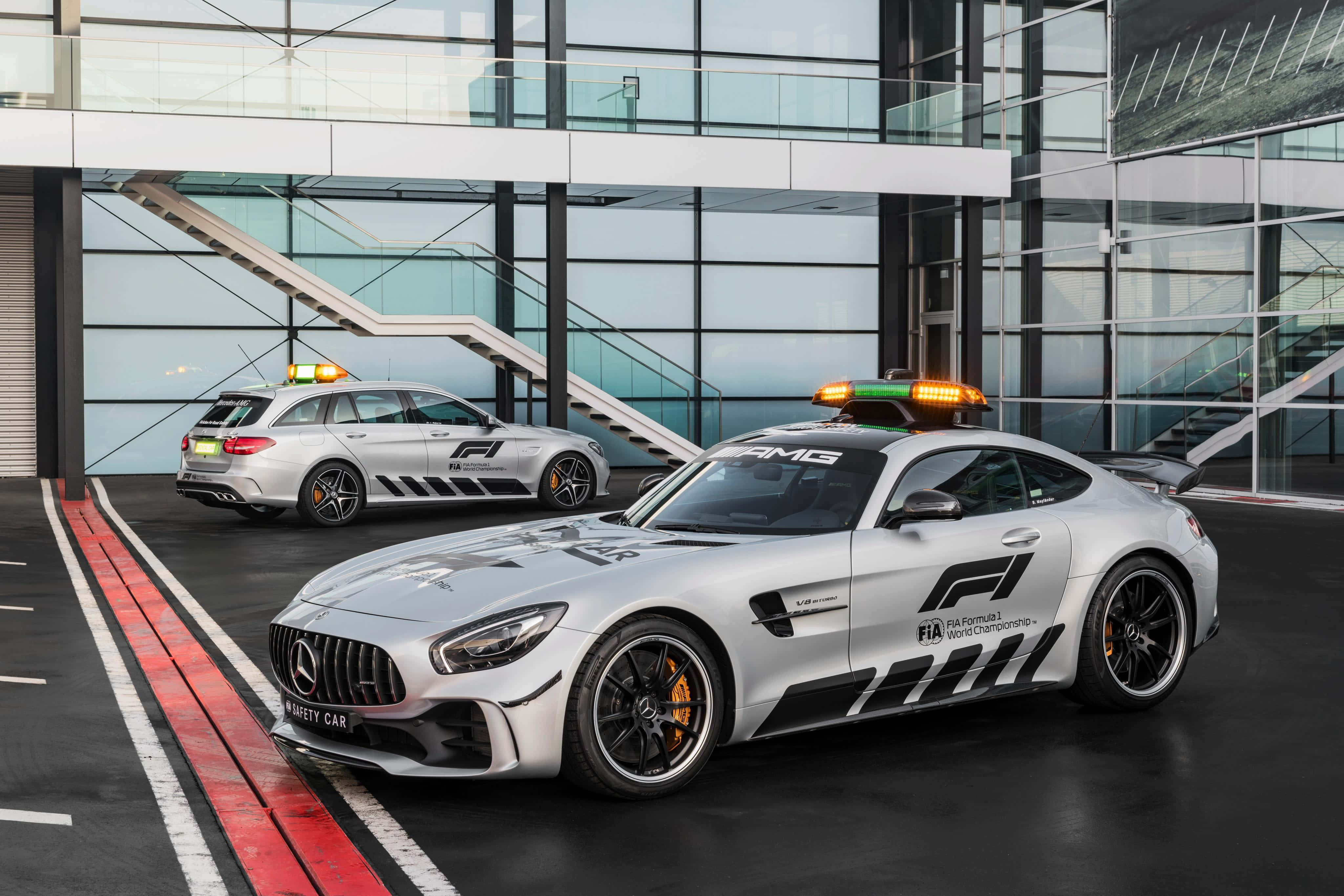Safety Car 桌布 壁紙 — 兩輛銀色 Mercedes-AMG 安全車，閃著燈，停在一棟現代玻璃建築前。