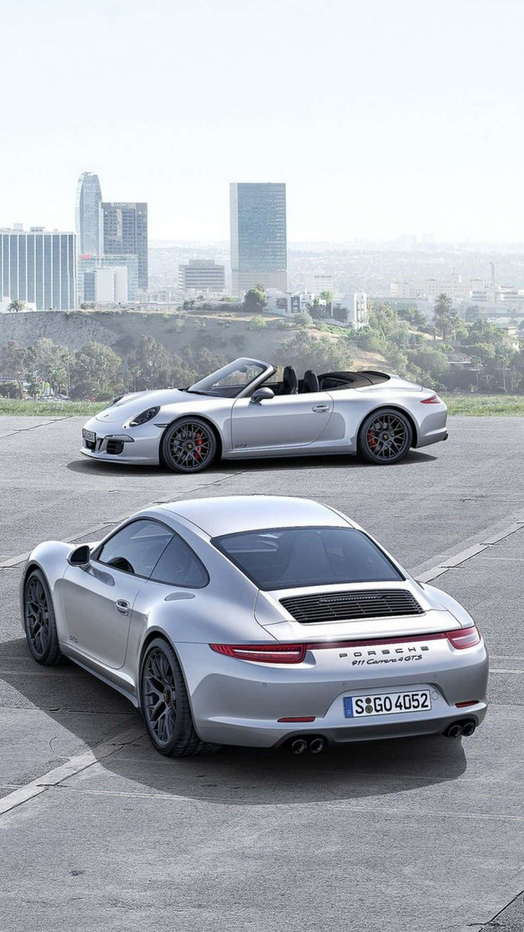 Porsche iPhone 桌布 壁紙 — 兩輛銀色保時捷911 Carrera GTS跑車停放在混凝土觀景台上，背景是朦朧的城市天際線。